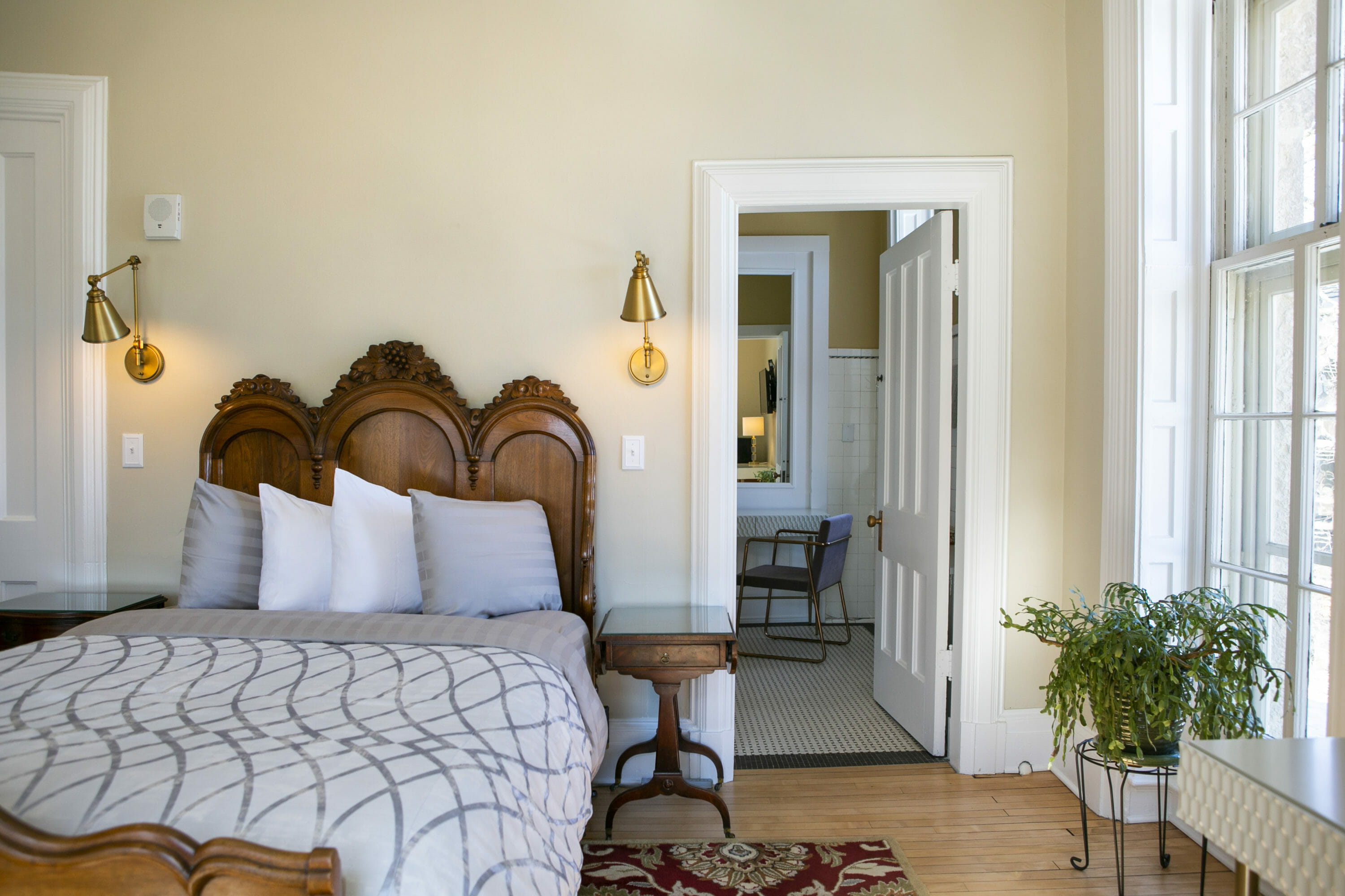 Cozy bedroom with carved wood headboard, brass sconces, and a view into an adjoining bathroom featuring vintage tile and a modern chair.