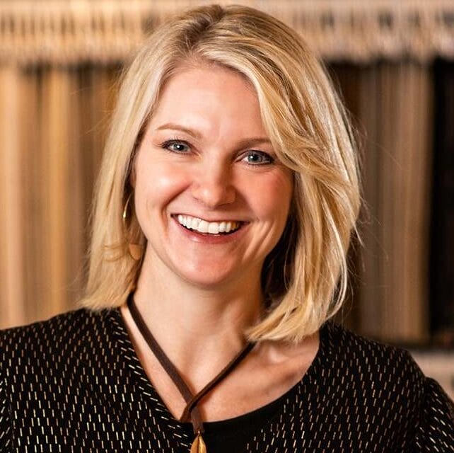 Smiling blonde woman with blue eyes wearing a patterned top and necklace.
