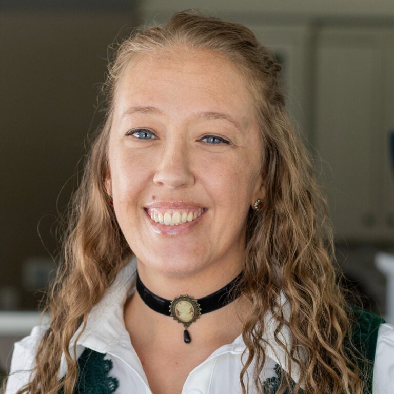 Woman with blue eyes and blonde hair smiles, wearing a black cameo choker and white shirt.