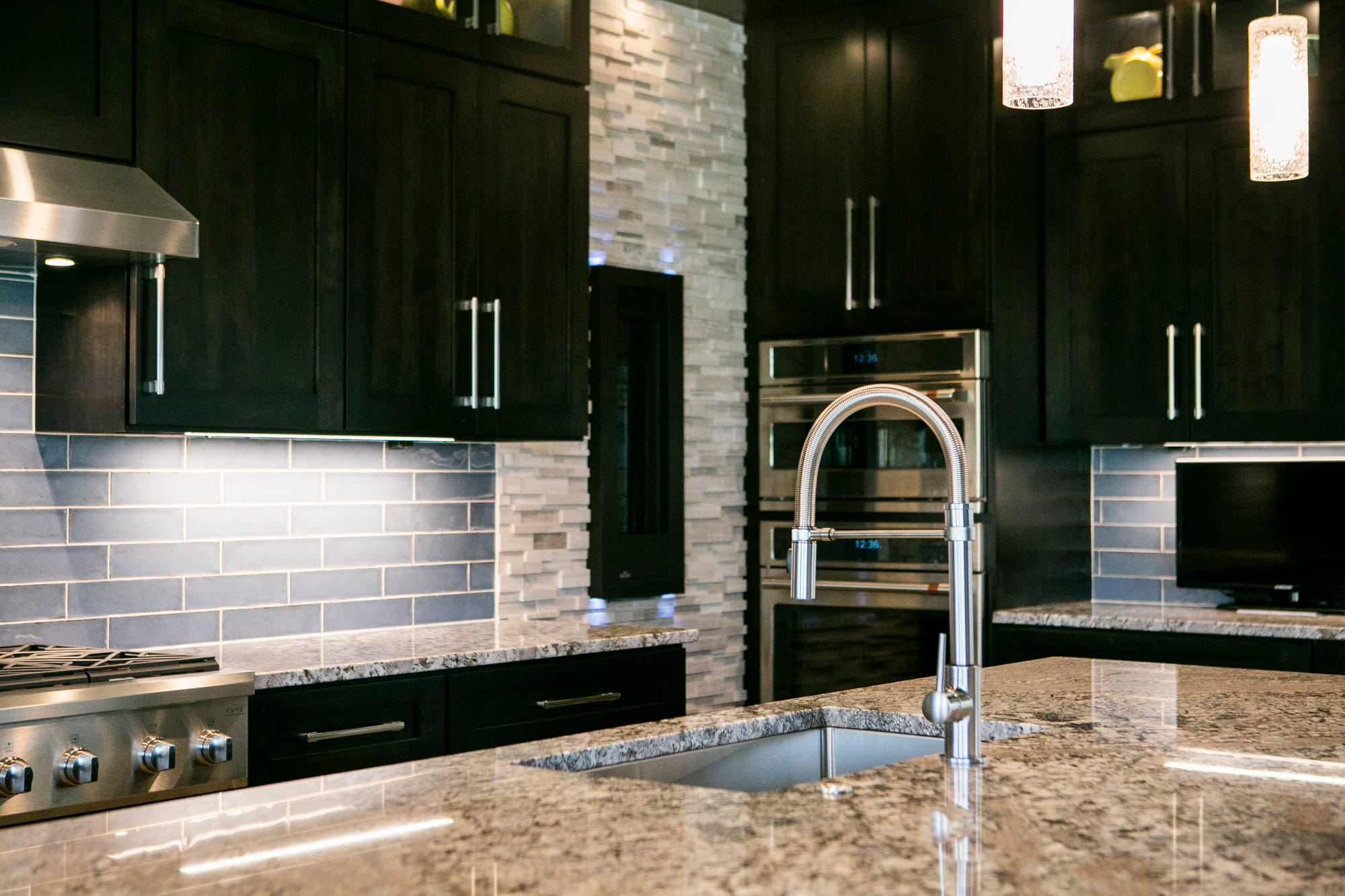 A stunning modern kitchen remodel in Madison, WI, featuring dark cabinetry, a sleek granite island with a stainless steel high-arc faucet, a stylish subway tile backsplash, and built-in double ovens. The design blends elegance and functionality with under-cabinet lighting and high-end appliances. Perfect for homeowners seeking luxury kitchen renovations and expert home remodeling. Contact top interior designers and home remodeling contractors in Madison, WI, for custom kitchen upgrades.