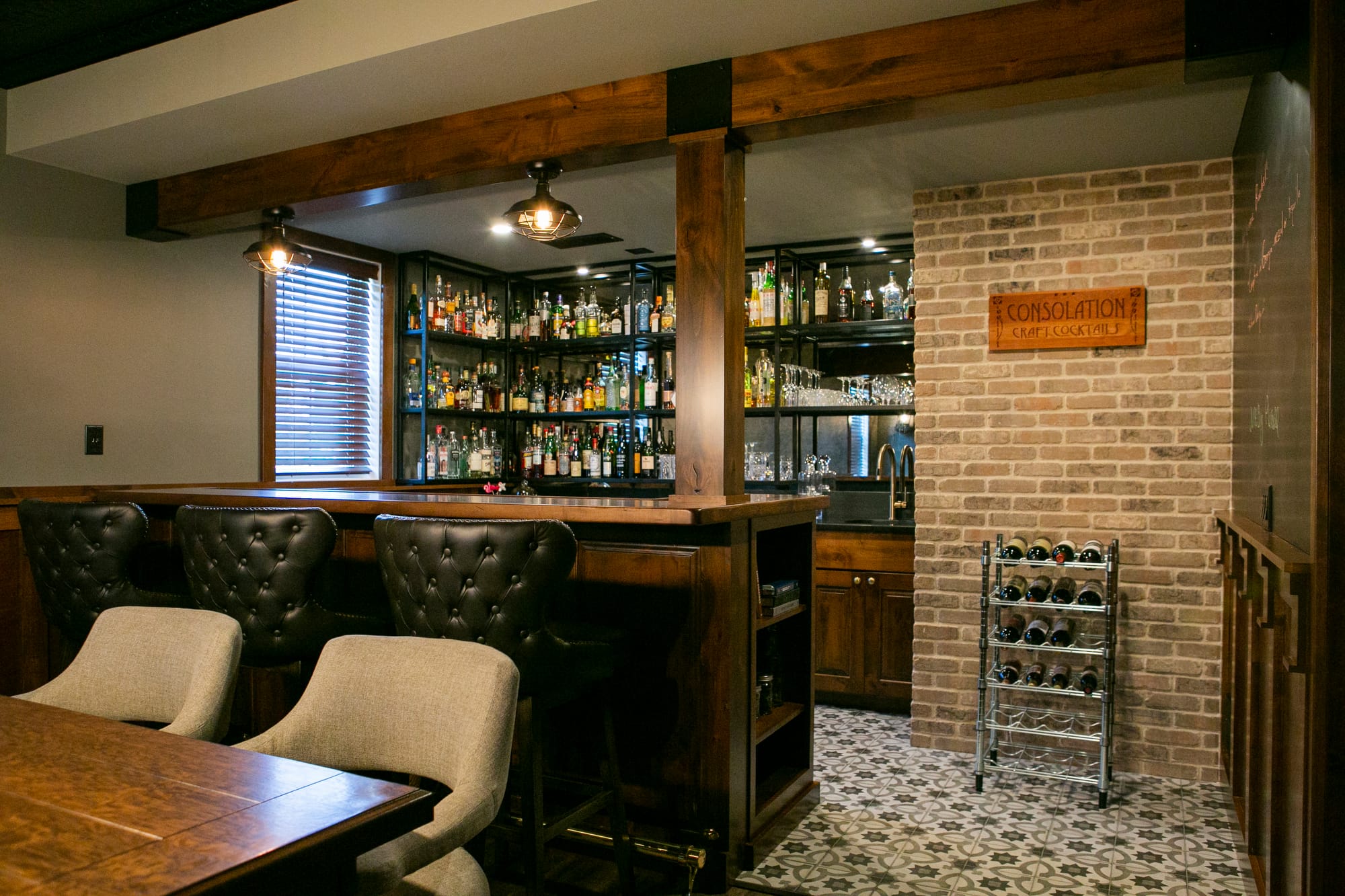 An elegant home bar and dining area featuring a rustic wooden bar with tufted black leather bar stools, complemented by a modern wooden dining table with gray upholstered chairs. The space is illuminated by an industrial-style light fixture with exposed Edison bulbs, creating a warm and inviting atmosphere. Perfect for home remodeling inspiration in Madison WI, showcasing a blend of classic and contemporary interior design.