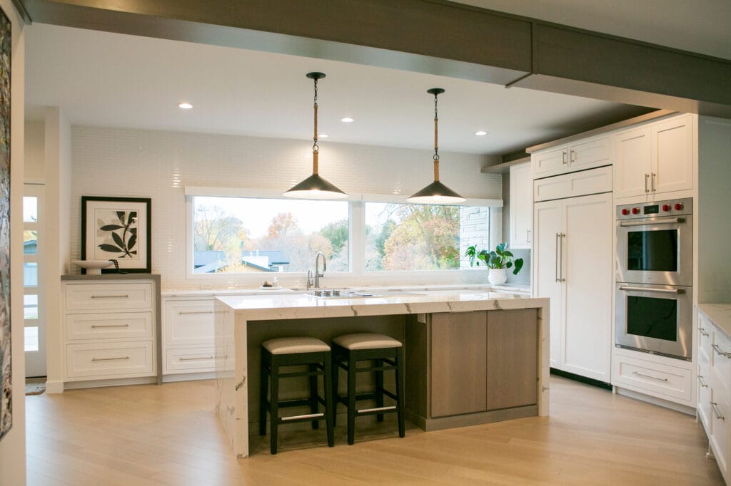 A spacious and modern white kitchen featuring a large marble waterfall island with two upholstered barstools. The space is illuminated by two black pendant lights with rope detailing, hanging over the island. White shaker-style cabinets contrast with light hardwood flooring, while stainless steel double ovens and a built-in refrigerator add a sleek, functional touch. A large window above the sink provides abundant natural light and a view of the autumn landscape outside. A potted plant and framed botanical artwork add warmth and elegance to the design.
