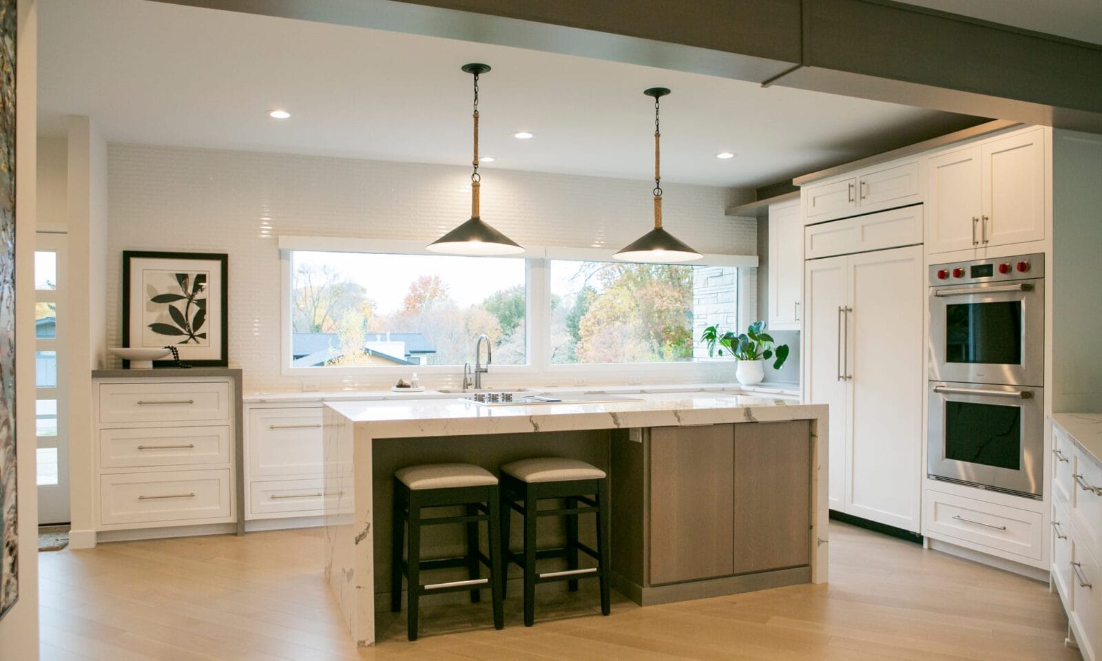 A spacious and modern white kitchen featuring a large marble waterfall island with two upholstered barstools. The space is illuminated by two black pendant lights with rope detailing, hanging over the island. White shaker-style cabinets contrast with light hardwood flooring, while stainless steel double ovens and a built-in refrigerator add a sleek, functional touch. A large window above the sink provides abundant natural light and a view of the autumn landscape outside. A potted plant and framed botanical artwork add warmth and elegance to the design.
