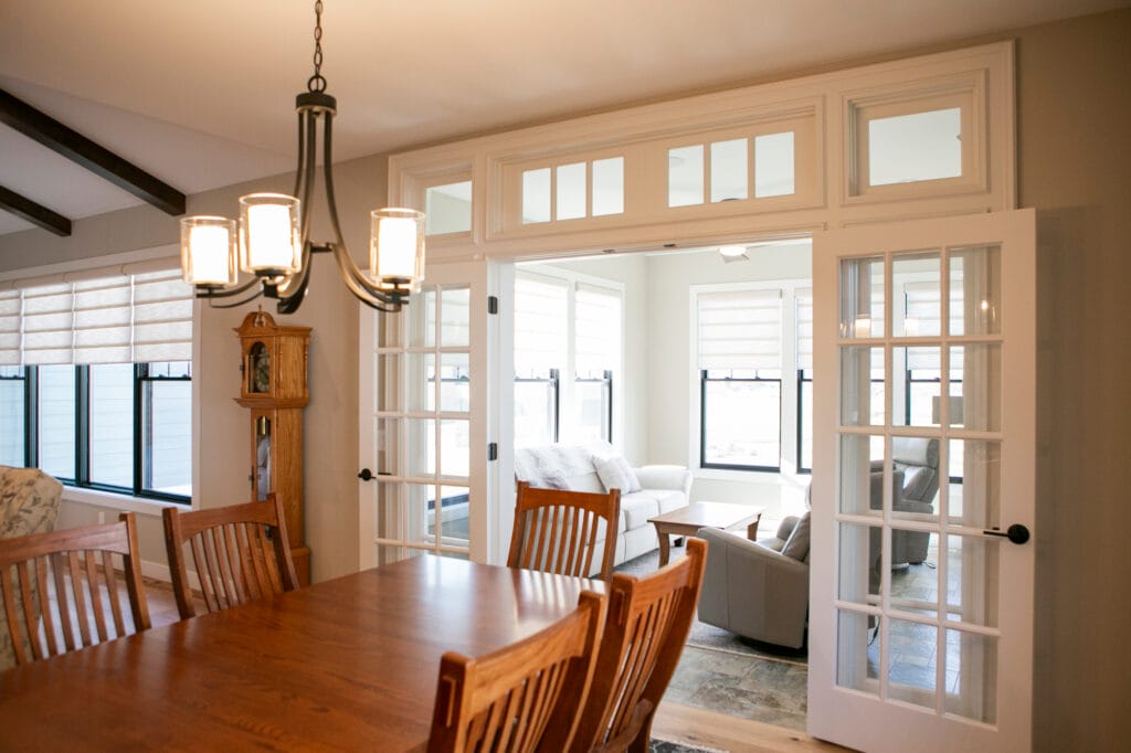 A beautifully designed open-concept dining room in Madison, WI featuring a rich wooden dining table with matching chairs, a stylish black chandelier, and large French doors leading to a bright sunroom with cozy seating. The space is enhanced with natural light from expansive windows and modern shades, making it a perfect inspiration for interior design, home remodeling, and renovation projects.