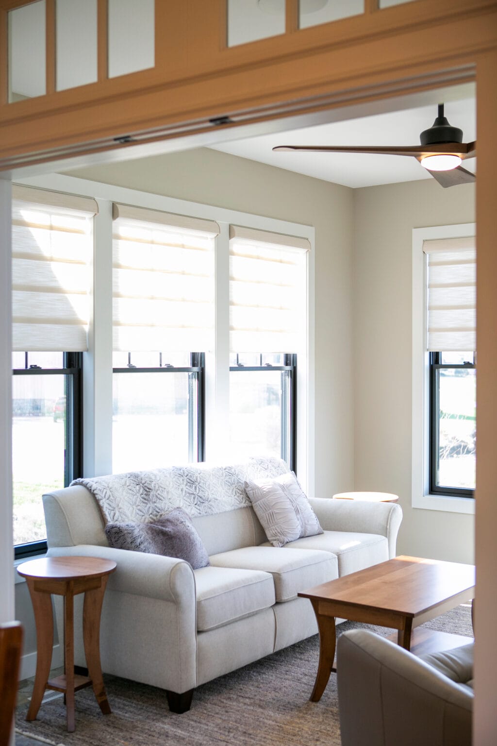 A beautifully lit sunroom featuring large black-framed windows with soft beige Roman shades, a plush cream-colored loveseat adorned with textured throw pillows, and a warm wooden coffee table. A modern ceiling fan with wood accents hangs above, enhancing the contemporary farmhouse aesthetic. The space is framed by elegant French doors with transom windows, creating an inviting and airy atmosphere.