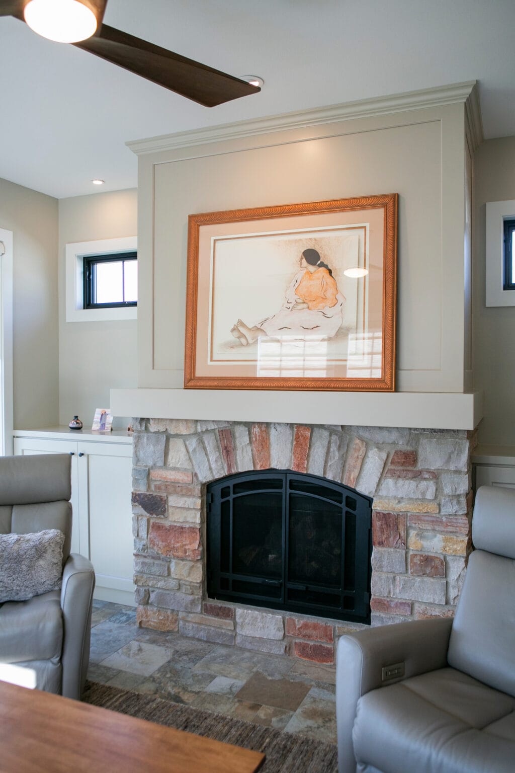 A beautifully designed living area featuring a stone fireplace with a white wood mantel, adorned with a framed artwork. Two sleek gray leather recliners with plush accent pillows sit in front of the fireplace, creating a cozy and inviting ambiance. Built-in cabinets with decorative accents enhance the modern yet warm aesthetic.
