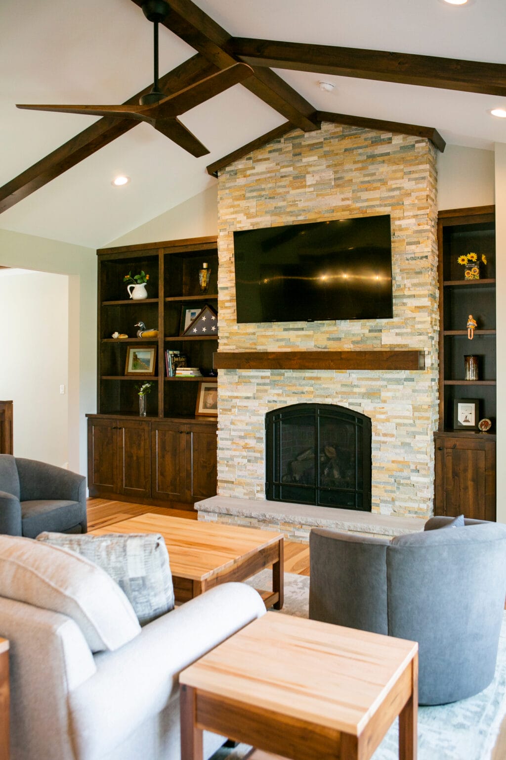 A beautifully designed living room in Madison, WI, featuring a stunning floor-to-ceiling stone fireplace with a wooden mantel, built-in dark wood cabinetry, and a mounted flat-screen TV. The space is complemented by exposed wood ceiling beams, modern furniture, and a neutral color palette. Designed by top interior designers in Madison, WI, this space offers inspiration for home remodeling, interior design, and modern living room renovations.