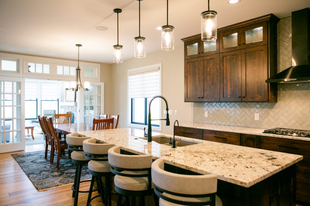 Elegant kitchen remodel in Madison, WI, featuring a large granite island with seating, custom dark wood cabinetry, modern pendant lighting, and a stylish herringbone tile backsplash. Designed by top interior designers and home remodeling contractors in Madison for a sophisticated and functional space.