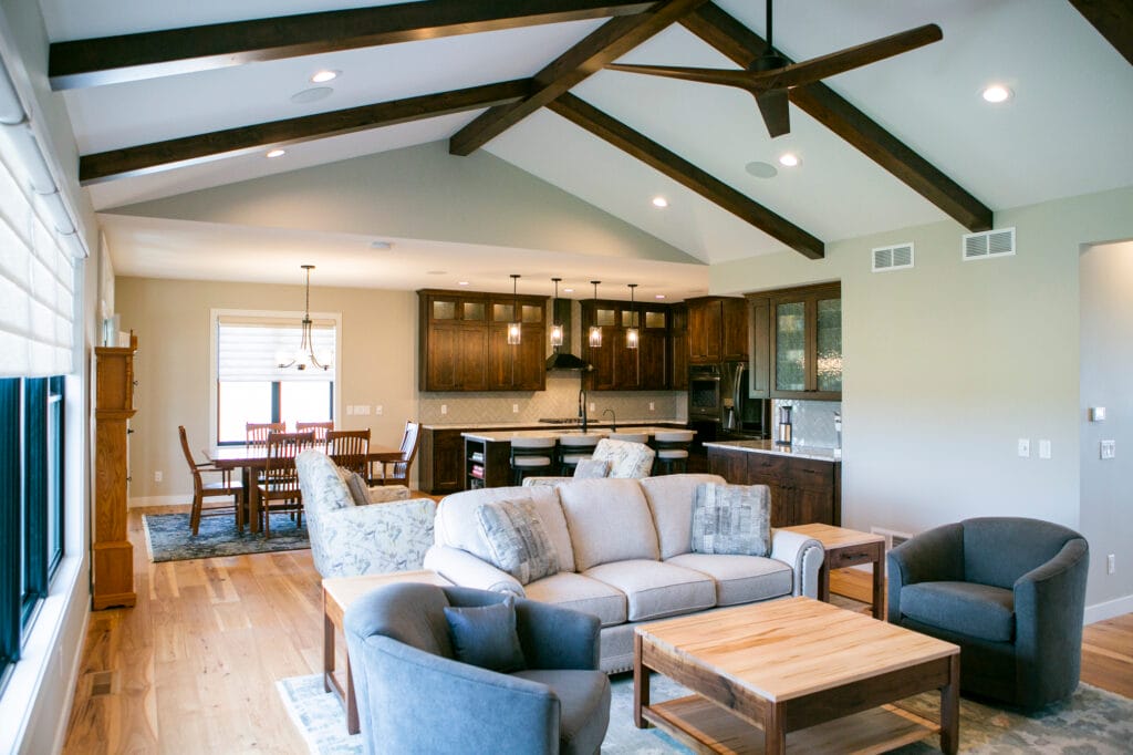 A beautifully designed open-concept living space featuring a vaulted ceiling with exposed beams, a modern ceiling fan, and a cozy seating area. The adjacent kitchen showcases dark wood cabinetry, pendant lighting, and a spacious island. Expertly crafted by leading interior designers in Madison, WI, this home remodel blends elegance with functionality.
