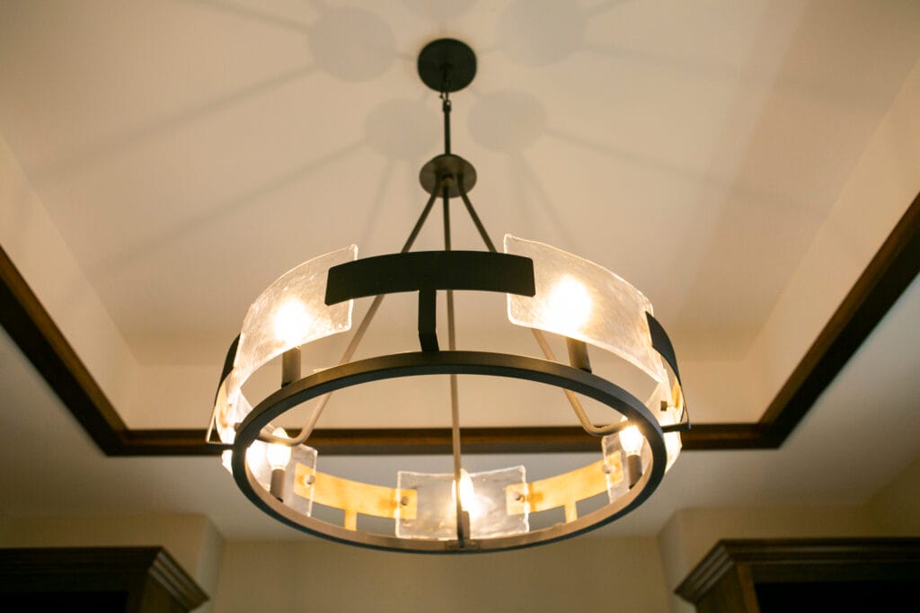 A close-up of a modern industrial-style chandelier featuring a circular black metal frame with frosted glass panels and warm lighting. The fixture hangs from the ceiling in an elegant room with dark wood trim, casting intricate shadows on the ceiling.