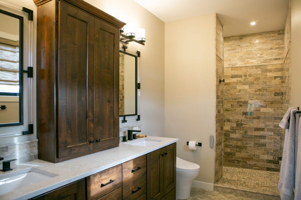 A beautifully designed bathroom renovation in Madison, WI, featuring elegant dark wood bathroom cabinets, a dual-sink vanity with quartz countertops, and modern black fixtures. The spacious walk-in shower is accented with natural stone tiles, creating a sophisticated and timeless interior. Designed by top interior designers in Madison, WI, this space showcases luxury home remodeling and high-end bathroom renovations.