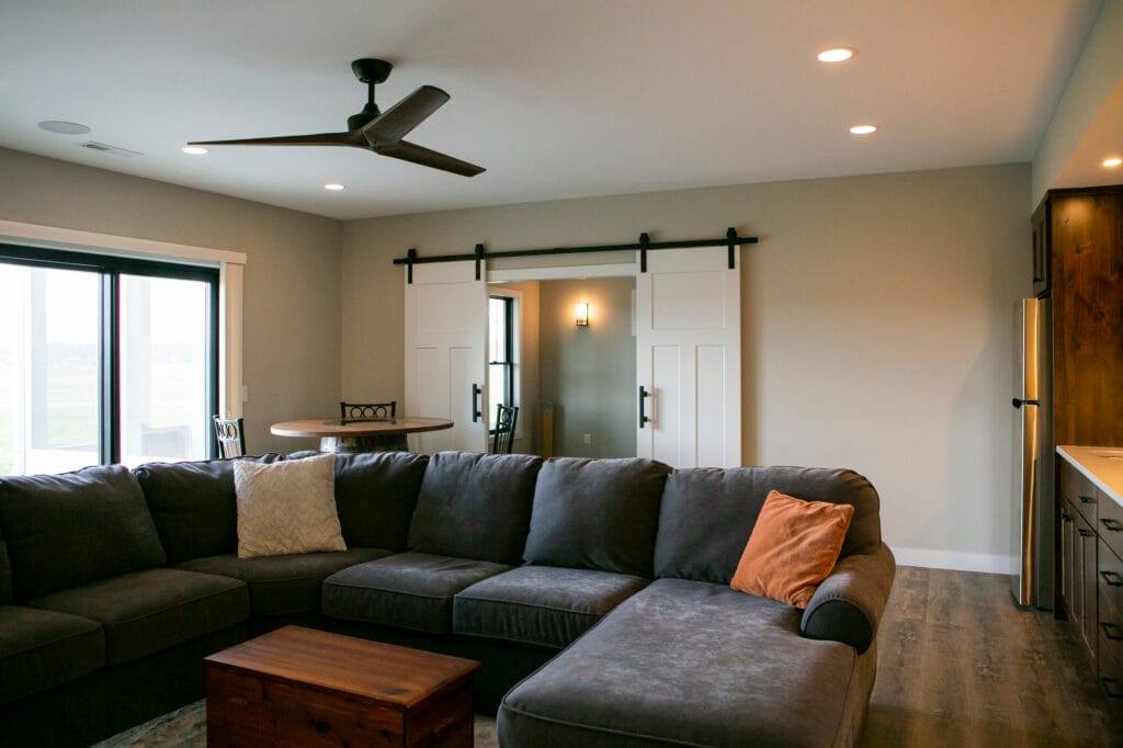 A stylish and modern living room in Madison, WI featuring a plush dark gray sectional, rustic wood coffee table, and elegant barn doors leading to an adjoining space. The open-concept design includes a small dining area and a kitchenette with stainless steel appliances, perfect for home renovation and remodeling inspiration.