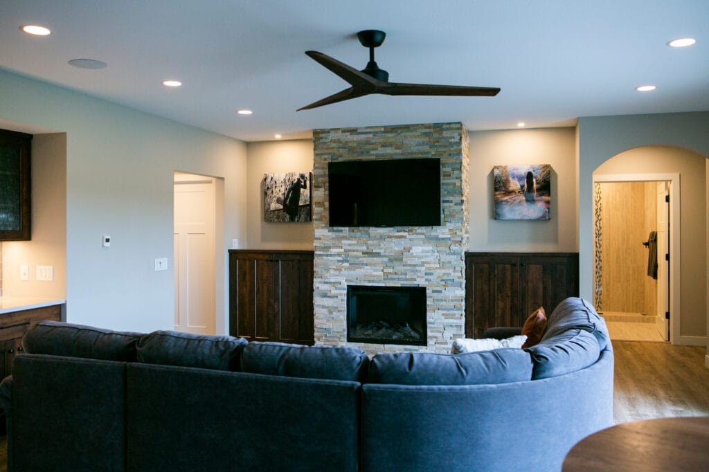 A beautifully designed living room in Madison, WI, featuring a modern stone fireplace with a mounted flat-screen TV, rich wood cabinetry, and a stylish ceiling fan. The open-concept space flows seamlessly into the kitchen, showcasing a luxurious home remodel with a cozy sectional sofa and warm lighting. Designed by top interior designers in Madison, WI, this home highlights expert interior design and home renovation excellence.