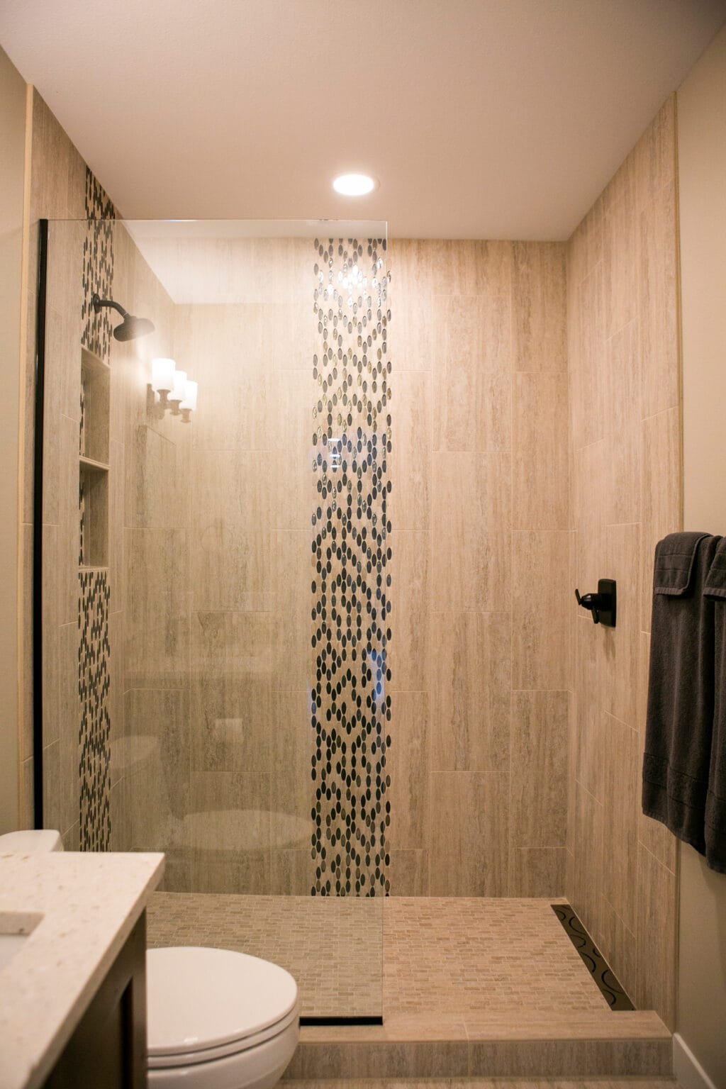 A beautifully renovated bathroom featuring a walk-in shower with a glass enclosure, beige textured tiles, and a striking black mosaic accent design. This modern bathroom remodel in Madison, WI, showcases high-end finishes, stylish lighting, and sleek black fixtures, making it a perfect blend of elegance and functionality. Designed by expert interior designers specializing in home renovations and bathroom remodels in Madison, WI.