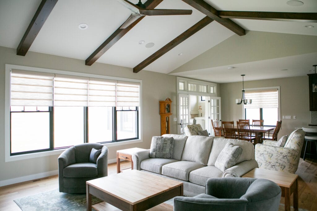 A spacious living room in Madison, WI featuring vaulted ceilings with exposed wooden beams, large windows with modern shades, and a cozy seating area with neutral-toned sofas and armchairs. The open-concept design flows into a dining space with a wooden dining table, making it a perfect inspiration for home remodeling and interior design projects.
