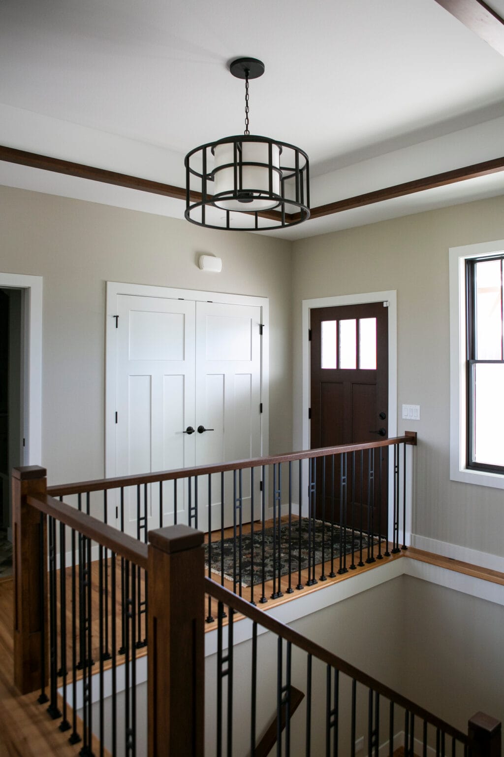 A stylish modern farmhouse entryway featuring a black metal and glass chandelier hanging from a white ceiling with dark wood beams. The space includes a dark wooden front door with glass panels, white double closet doors, and a staircase with a wood and black metal railing. Natural light streams through a large window, enhancing the neutral-toned walls and hardwood flooring.