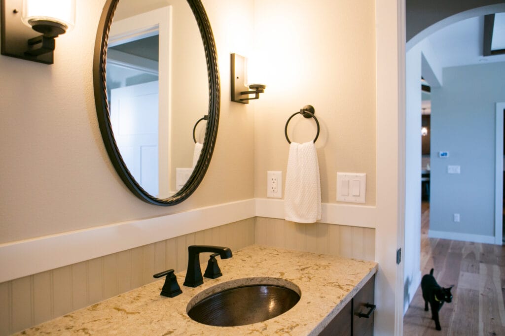 A beautifully designed powder room featuring a round black-framed mirror, warm lighting, a granite countertop with a copper sink, and black matte fixtures. The beadboard wainscoting adds a farmhouse touch, while a black cat walks in the hallway, adding charm to the cozy home ambiance.