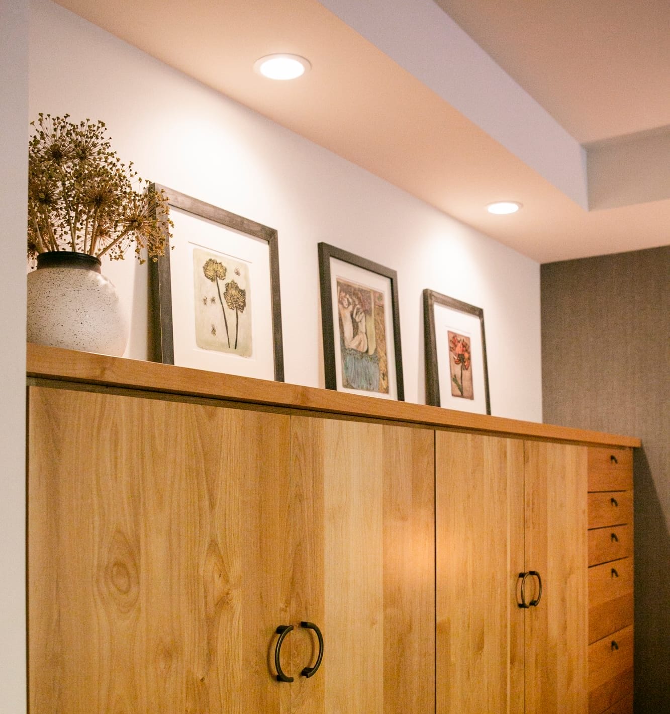 The cabinetry is crafted from light wood, featuring clean lines and a minimalist design. Two sets of double doors with black metal pull handles are positioned in the center, while a vertical row of smaller drawers with matching black hardware is located on the right side, adding both functionality and style. The top of the unit is styled as a display surface, adorned with framed artwork in black frames, each featuring unique botanical and artistic illustration.