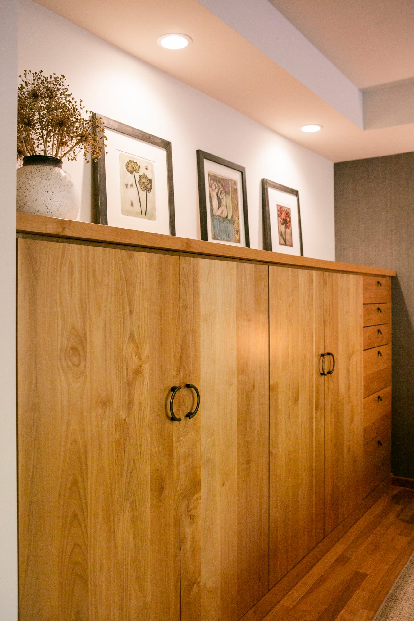The cabinetry is crafted from light wood, featuring clean lines and a minimalist design. Two sets of double doors with black metal pull handles are positioned in the center, while a vertical row of smaller drawers with matching black hardware is located on the right side, adding both functionality and style. The top of the unit is styled as a display surface, adorned with framed artwork in black frames, each featuring unique botanical and artistic illustration.