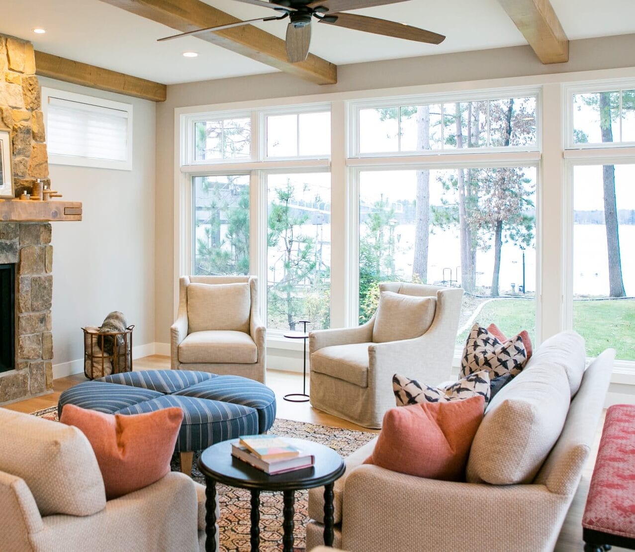 A warm and inviting living room with a focus on natural elements and ample light. The centerpiece is a beautiful stone fireplace with a rustic wooden mantle that holds decorative items, including a framed piece of artwork. Flanking the fireplace are two small rectangular windows with white blinds, letting in soft, diffused light. The ceiling features exposed wooden beams, adding warmth and a touch of rustic charm to the room. A ceiling fan with wooden blades provides a functional and stylish accent. The seating arrangement consists of two beige sofas and two plush armchairs in a neutral tone. The sofas are accented with soft throw pillows in muted shades of coral, navy, and patterned designs, adding a cozy and colorful touch. A striped blue ottoman in the center of the room serves as both a footrest and a casual table, surrounded by a patterned area rug that anchors the furniture.