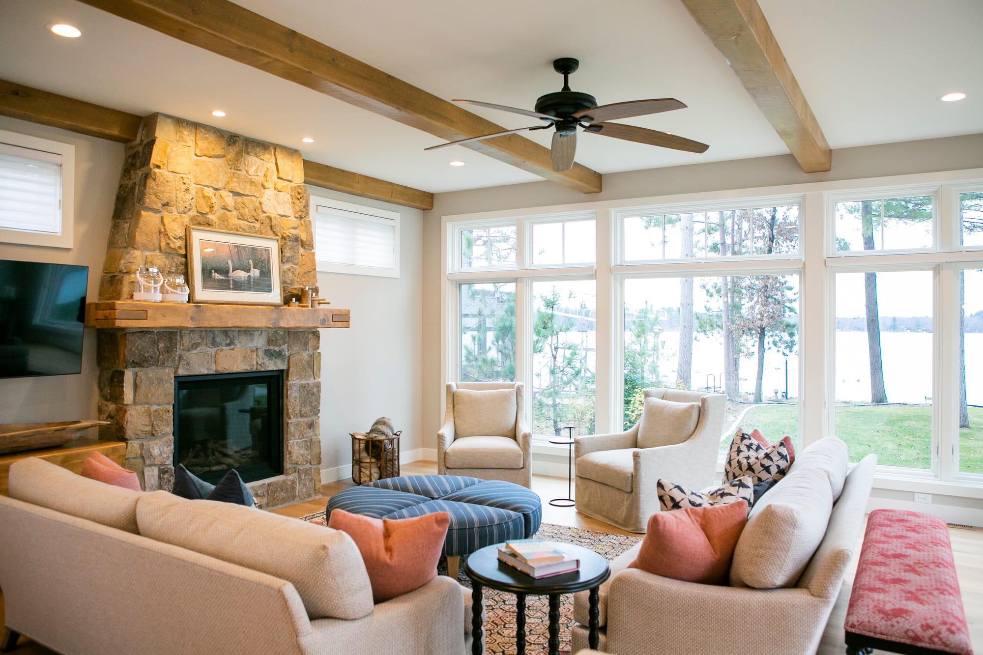 A warm and inviting living room with a focus on natural elements and ample light. The centerpiece is a beautiful stone fireplace with a rustic wooden mantle that holds decorative items, including a framed piece of artwork. Flanking the fireplace are two small rectangular windows with white blinds, letting in soft, diffused light. The ceiling features exposed wooden beams, adding warmth and a touch of rustic charm to the room. A ceiling fan with wooden blades provides a functional and stylish accent. The seating arrangement consists of two beige sofas and two plush armchairs in a neutral tone. The sofas are accented with soft throw pillows in muted shades of coral, navy, and patterned designs, adding a cozy and colorful touch. A striped blue ottoman in the center of the room serves as both a footrest and a casual table, surrounded by a patterned area rug that anchors the furniture.