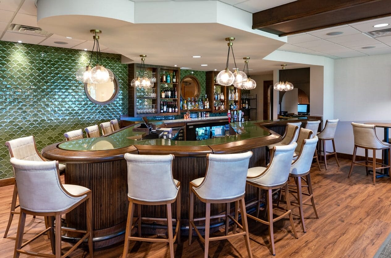 Art deco green and deep wooden bar. The shape of the bar is unique and is accompanied by white leather chairs. The bar backsplash is scalloped green.