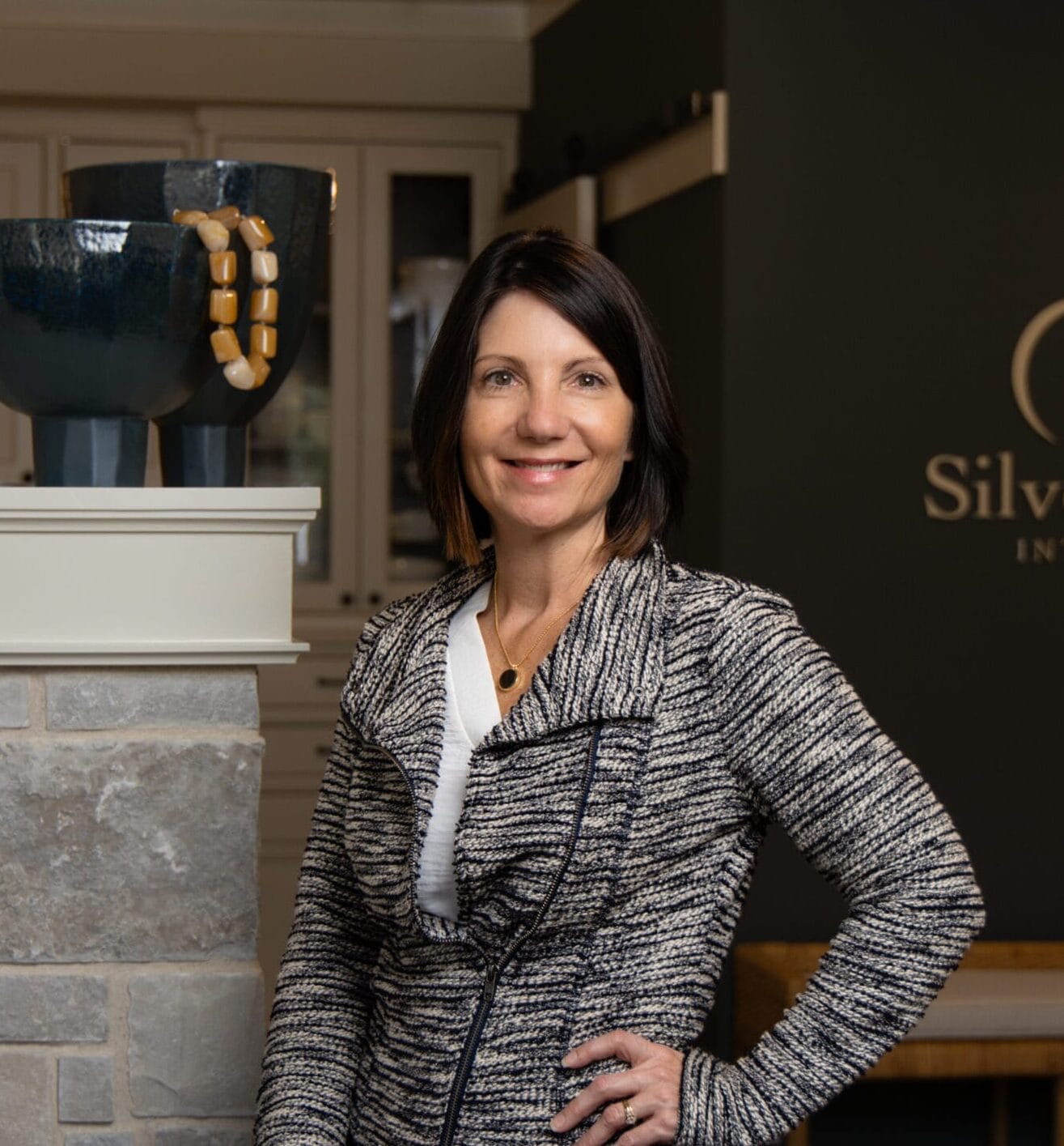 A woman with shoulder-length dark hair smiles, standing beside decorative bowls and a sign for Silverstone Interiors.