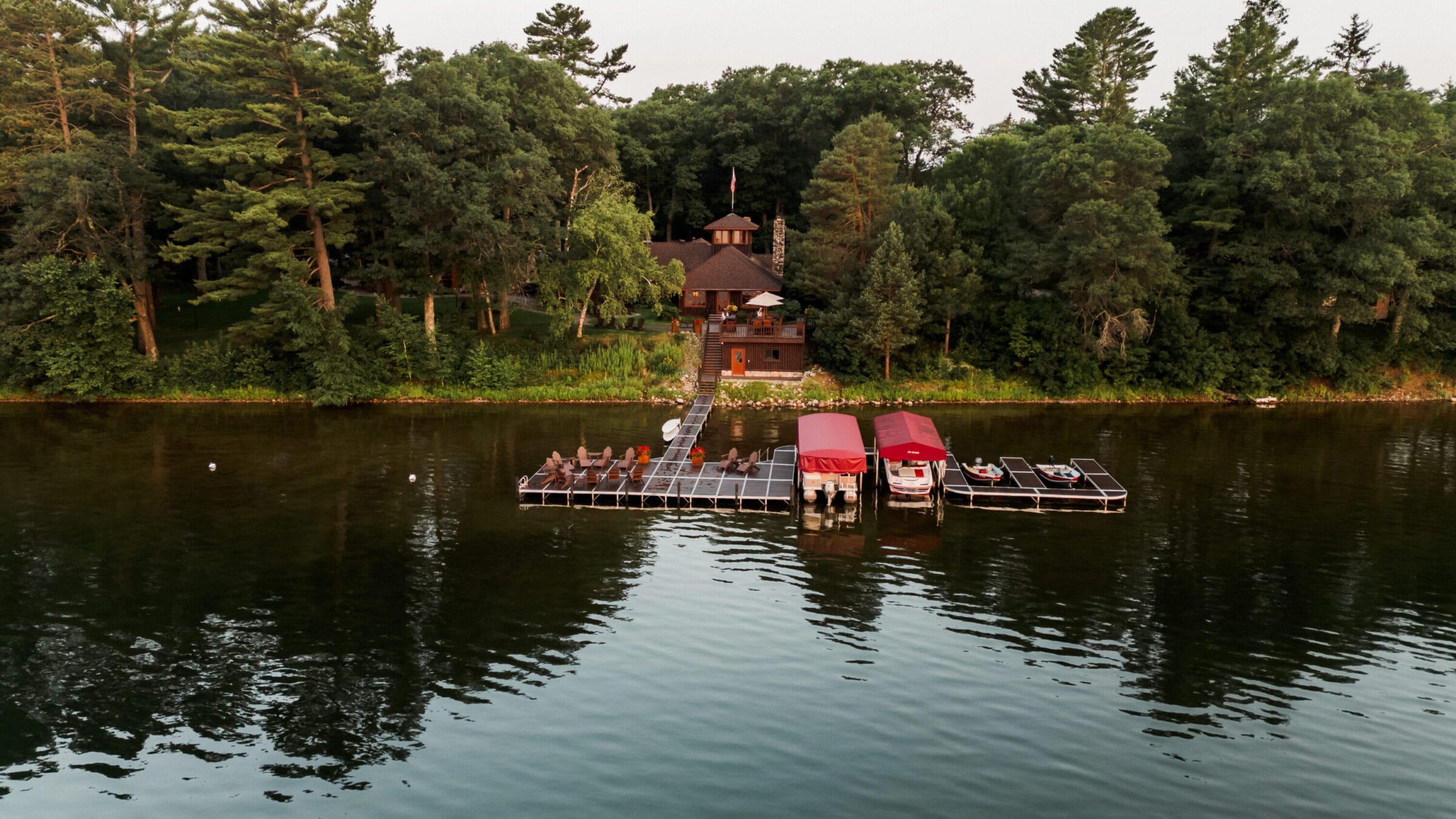Lakeside cabin surrounded by trees, featuring a dock with boats and seating, reflecting in calm water.