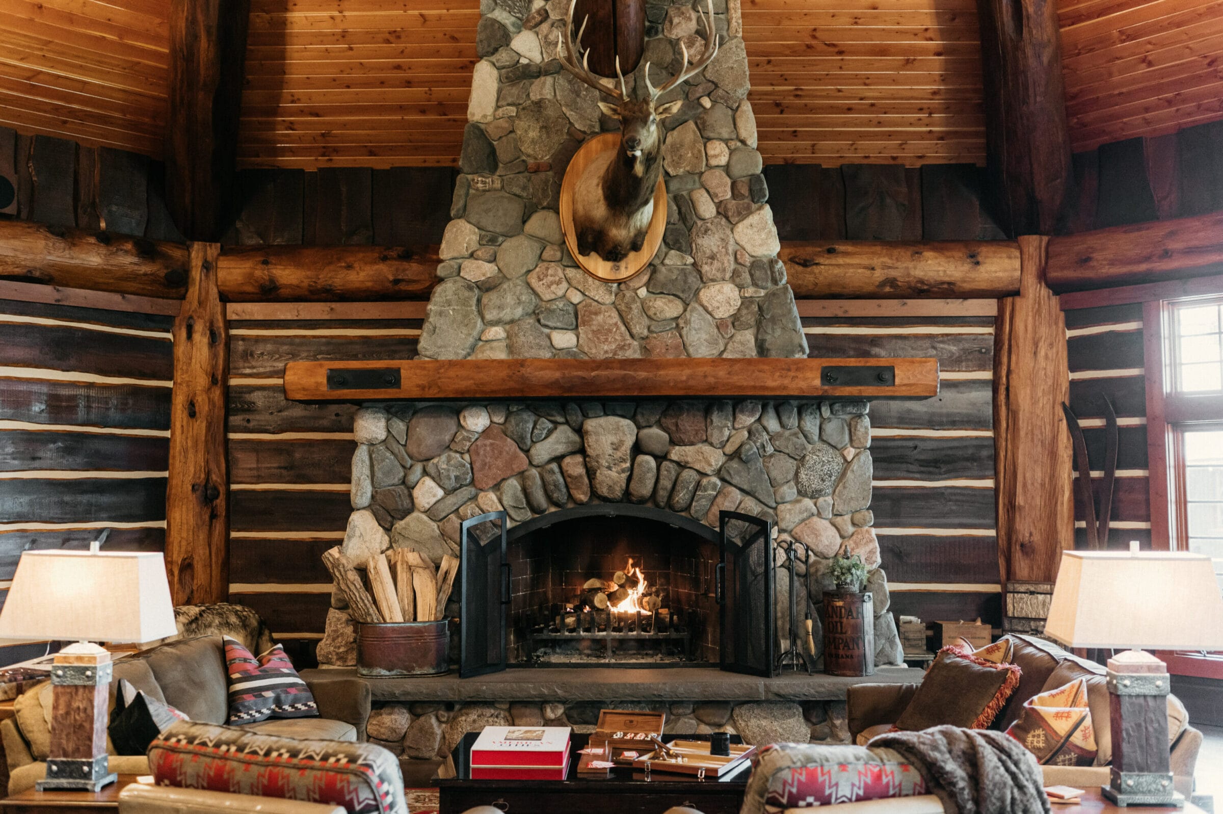 Cozy log cabin interior featuring a stone fireplace, wooden beams, and rustic decor with a mounted deer head.