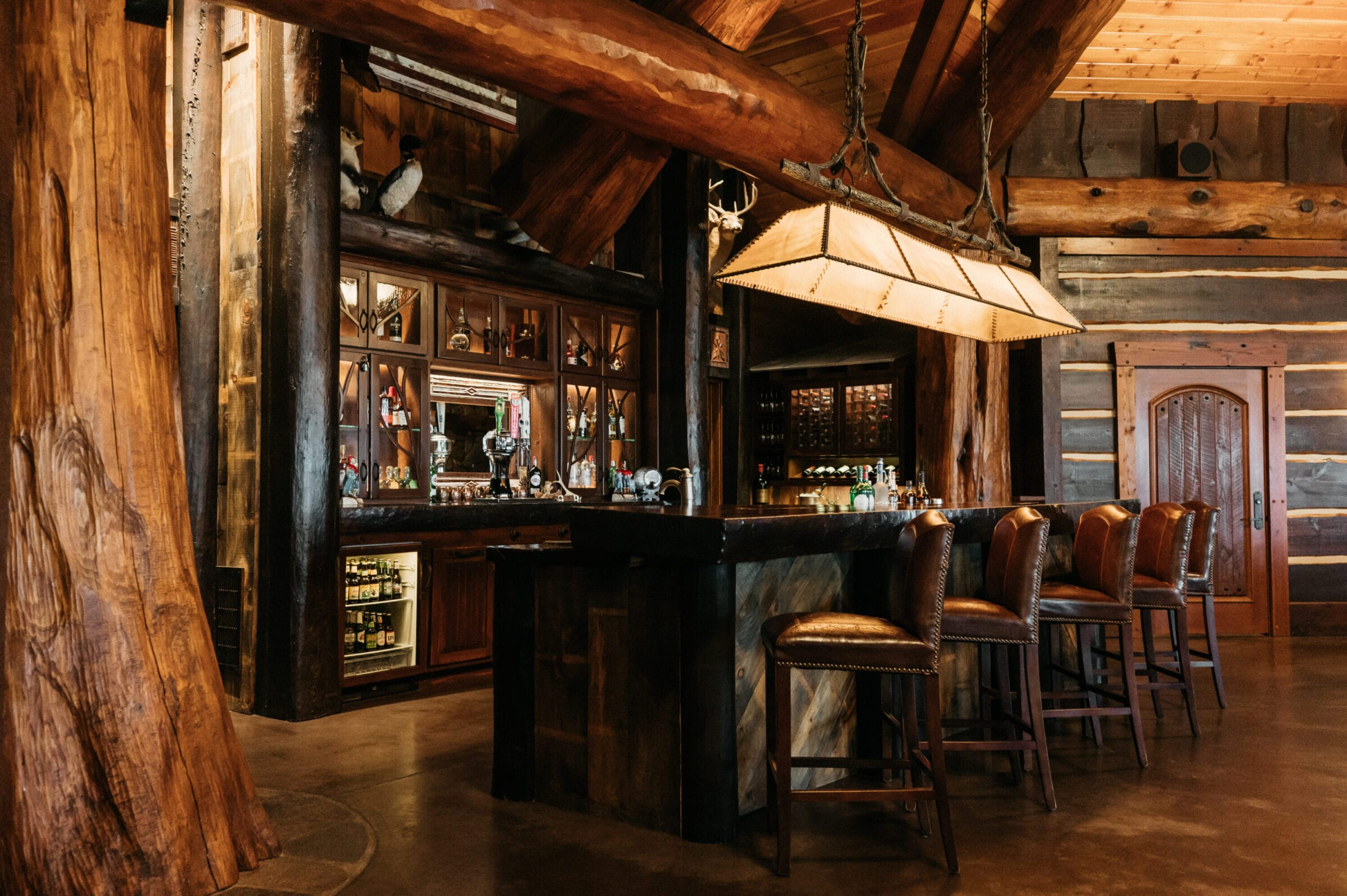 Rustic bar with wooden beams, leather stools, and a display of liquor bottles in a cozy, cabin-like setting.