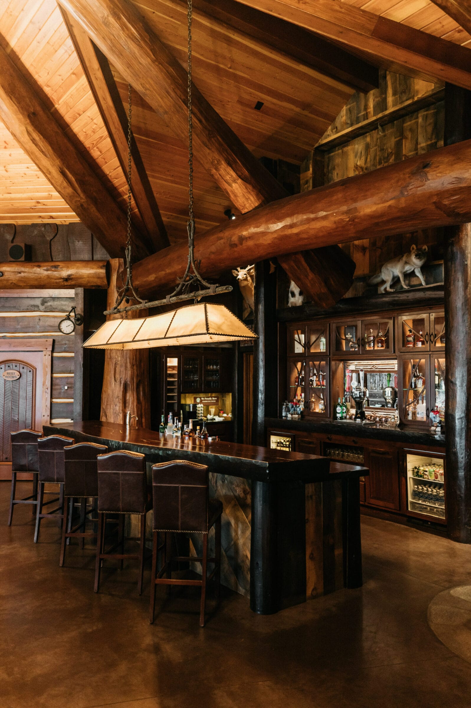 Rustic bar with leather stools, wooden beams, and a display of spirits in a cozy, lodge-style interior.