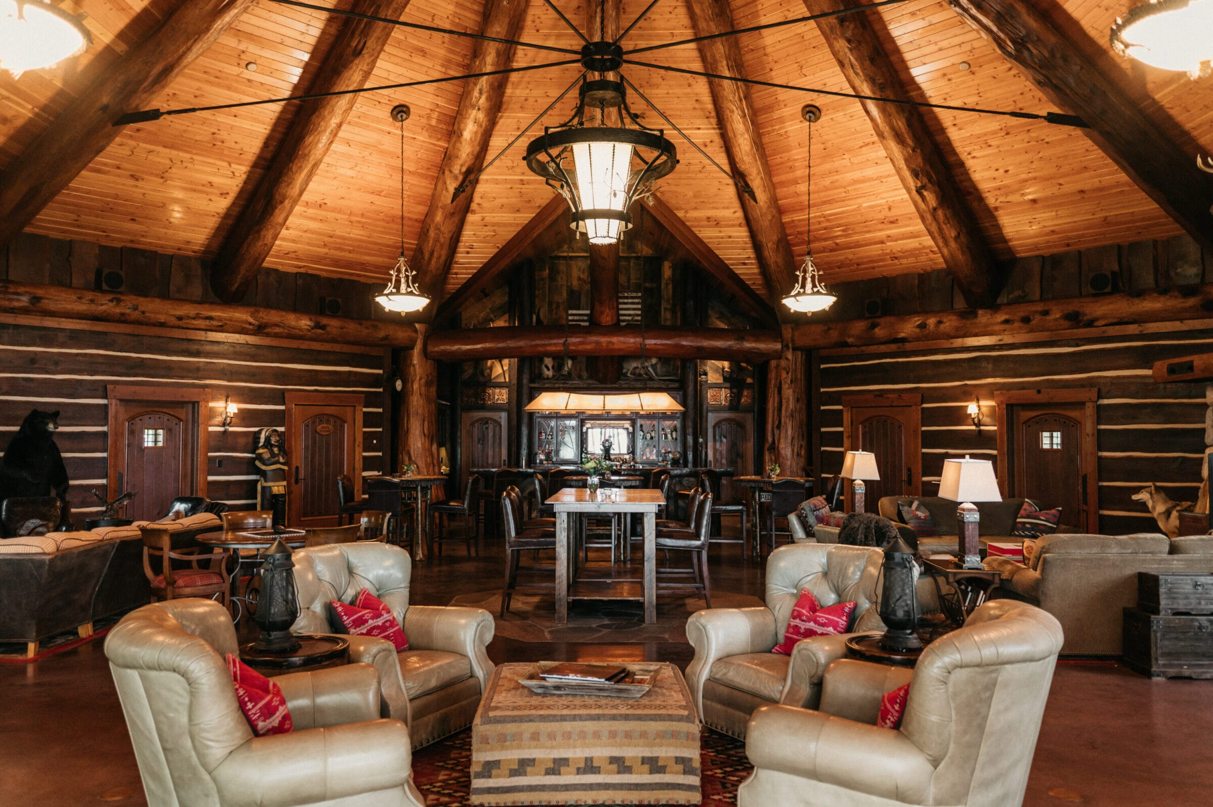 Rustic lodge interior featuring log walls, high wooden ceilings, cozy seating, and a central dining area with warm lighting.