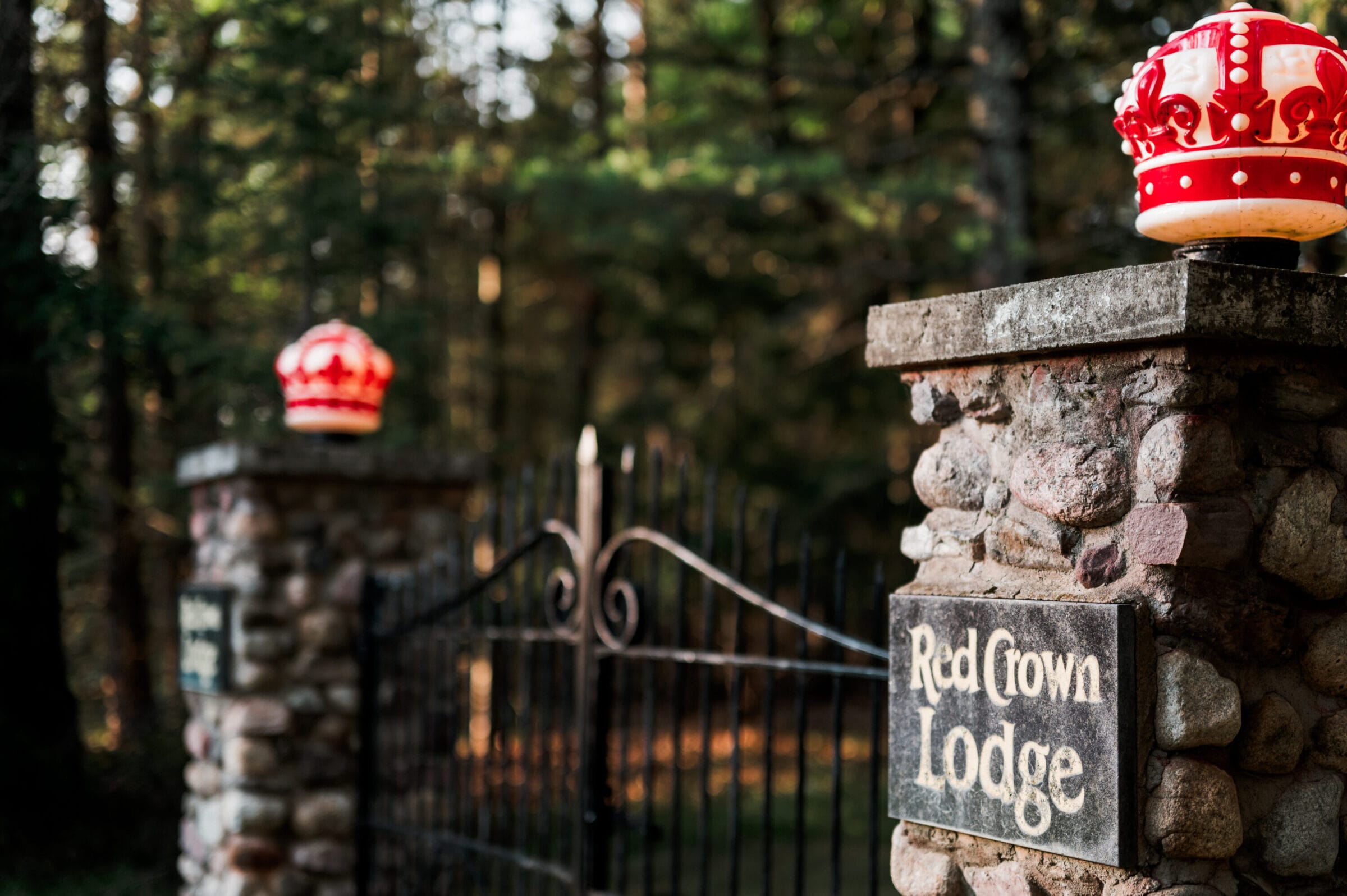 Stone gateposts with a sign reading "Red Crown Lodge" and red crowns atop, surrounded by a lush forest backdrop.