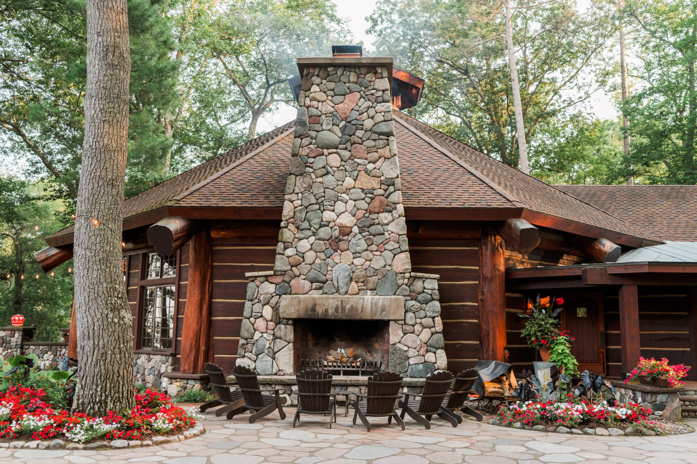 Rustic log cabin with a stone chimney, surrounded by lush trees and vibrant flower beds, featuring a cozy outdoor seating area.