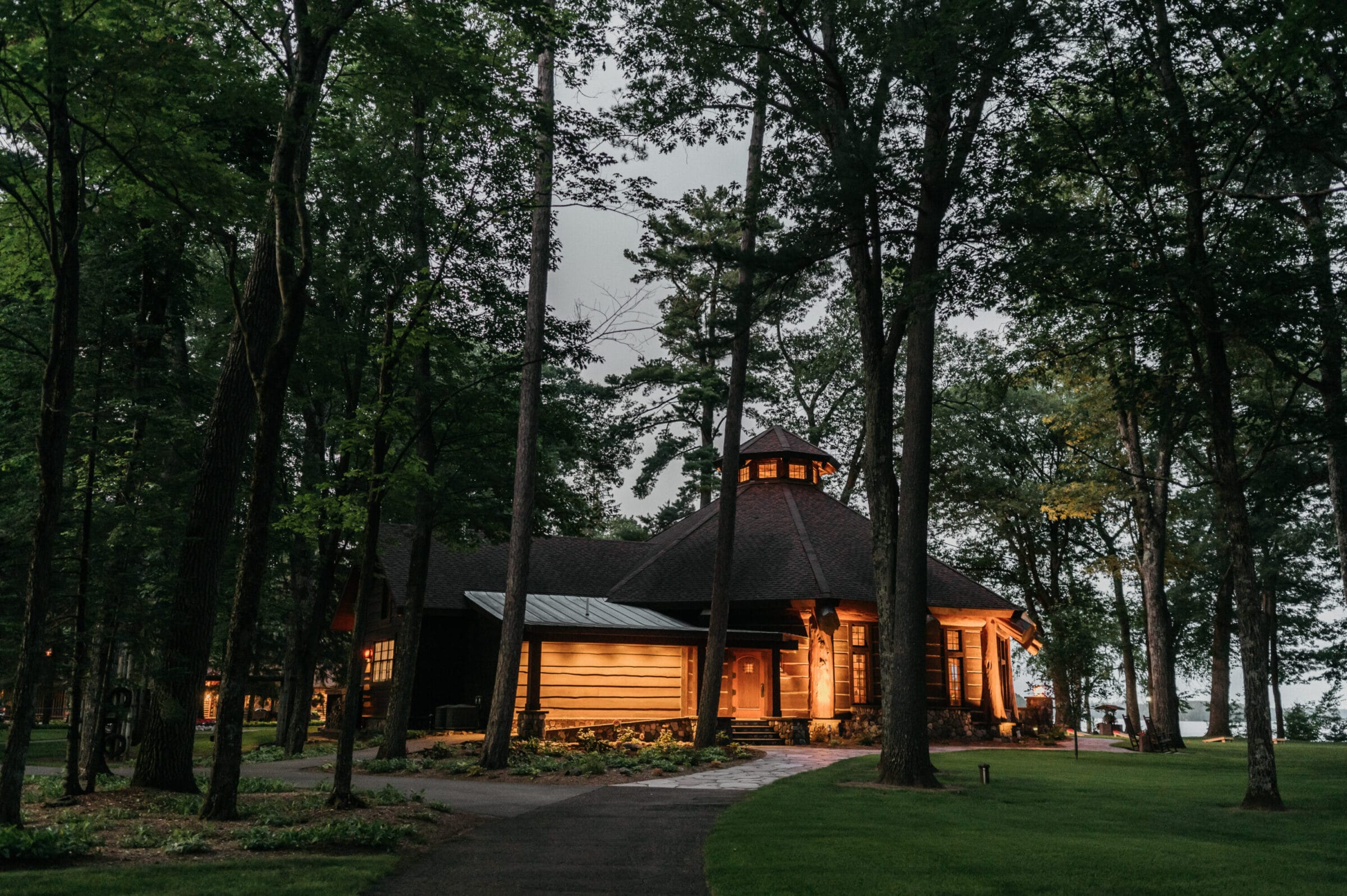 Cozy cabin nestled among tall trees, softly illuminated at dusk, with a winding path leading to its welcoming entrance.
