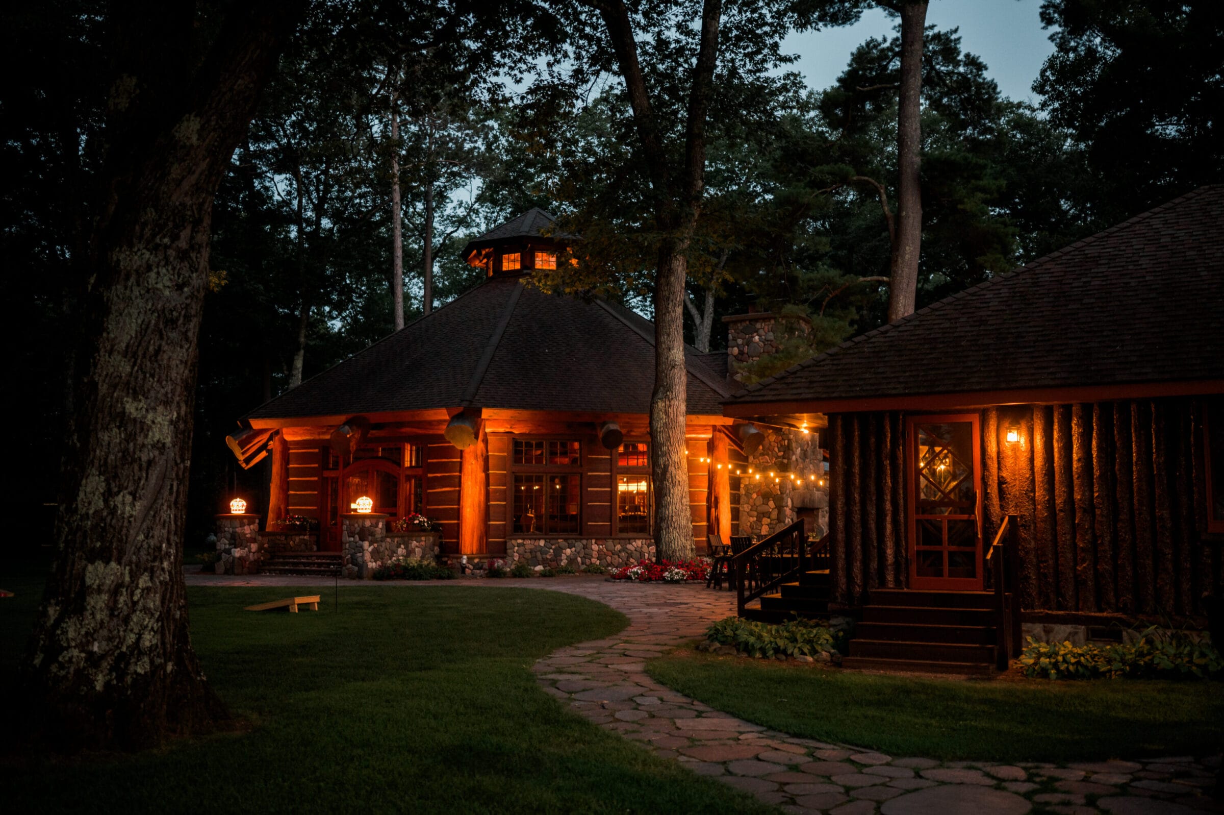 Cozy log cabin illuminated at dusk, surrounded by trees, with a winding stone path and warm outdoor lighting.