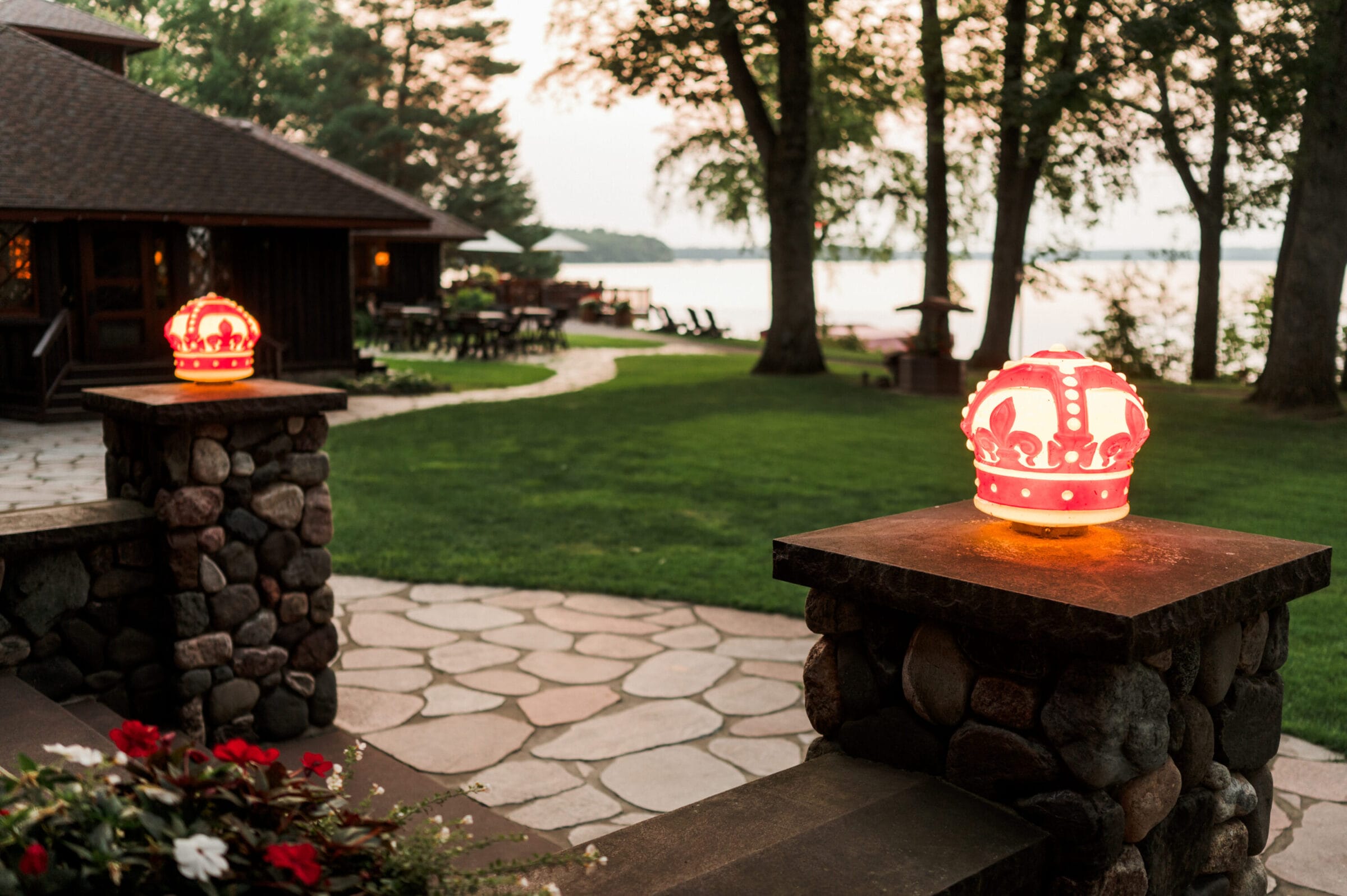 Glowing crown-shaped lanterns atop stone pillars, with a lush green lawn and lake in the background at sunset.