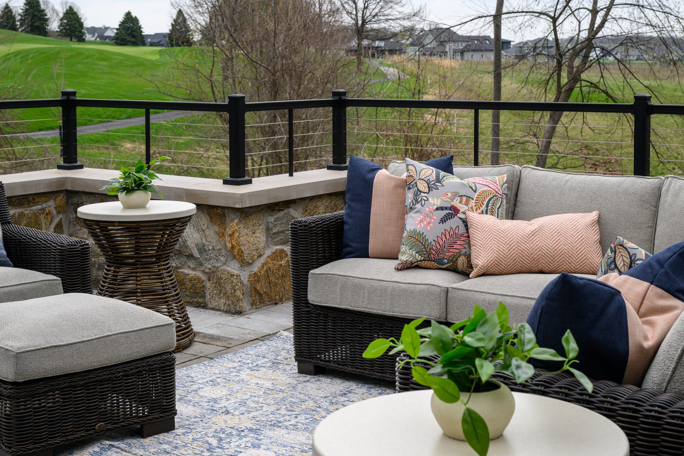 Outdoor patio seating area with woven wicker furniture, including a sofa and ottoman topped with neutral cushions and colorful patterned throw pillows. A small round side table with a potted plant sits beside the seating on a textured outdoor rug. The space is bordered by a low stone wall and black metal railing, overlooking a green lawn, leafless trees, and a quiet residential neighborhood in the distance under an overcast sky.