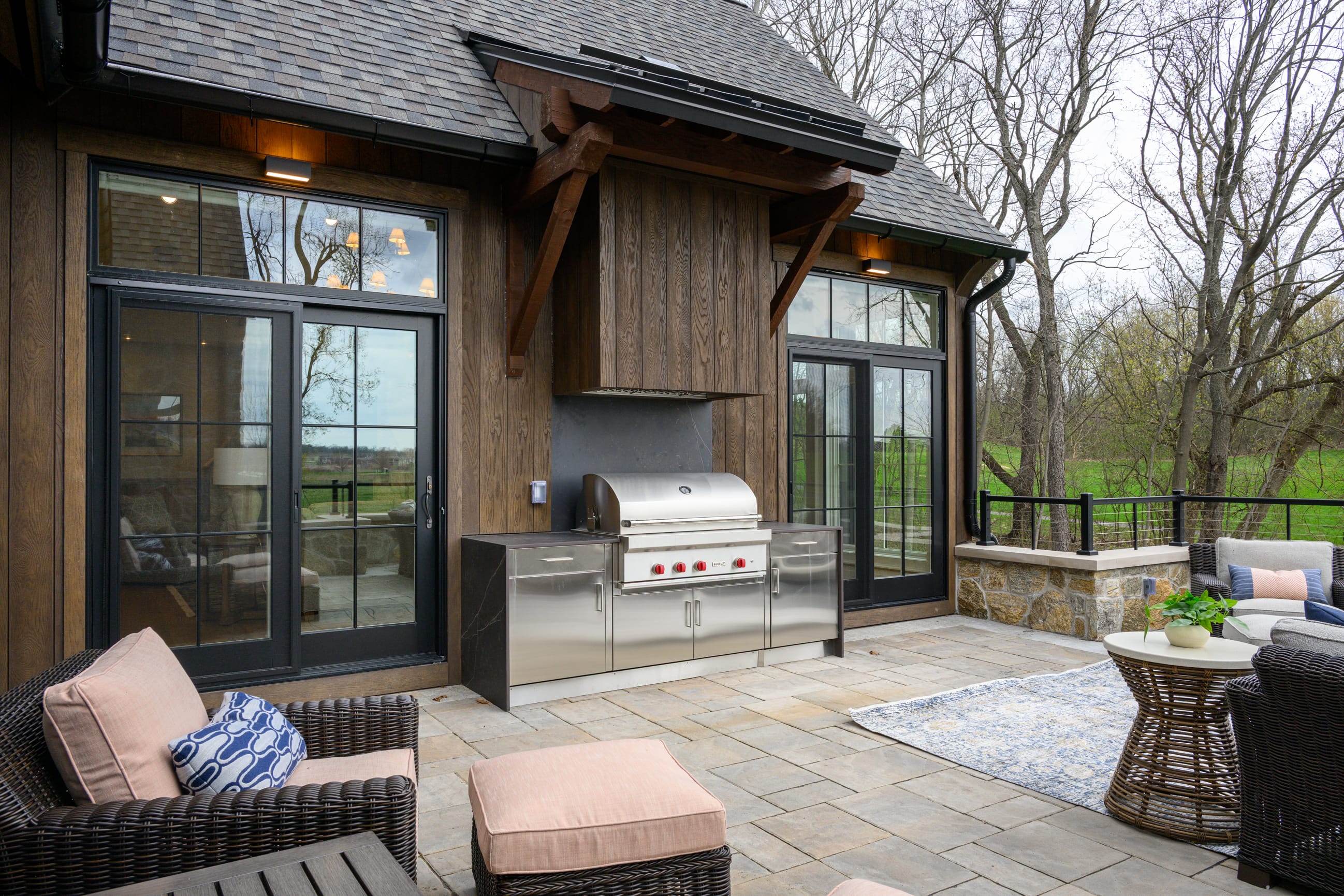 Stylish outdoor patio featuring a stainless steel grill, cozy seating, and large windows overlooking a green landscape.