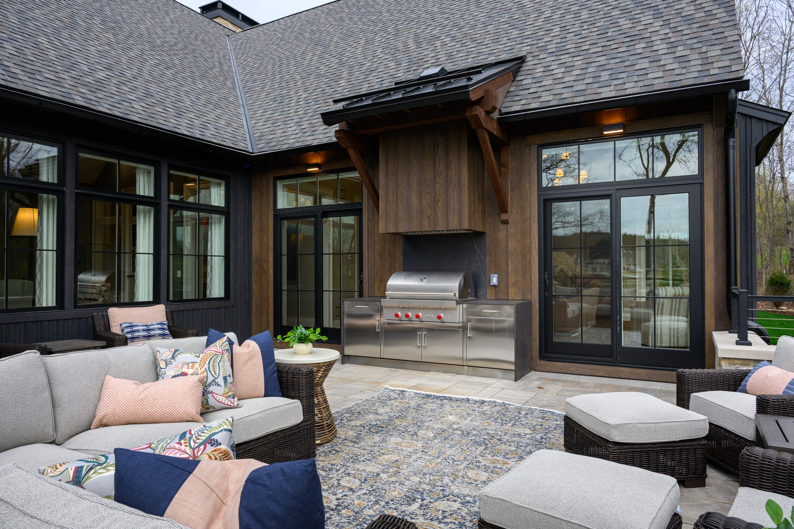 Outdoor patio attached to a modern farmhouse-style home, featuring a built-in stainless steel grill set into a stone and wood exterior wall beneath a small overhang. The patio includes woven wicker seating with neutral cushions and colorful patterned pillows arranged around a textured outdoor rug. Large black-framed windows and double glass doors line the house, reflecting nearby trees, while the space opens to a landscaped yard with leafless trees under an overcast sky.