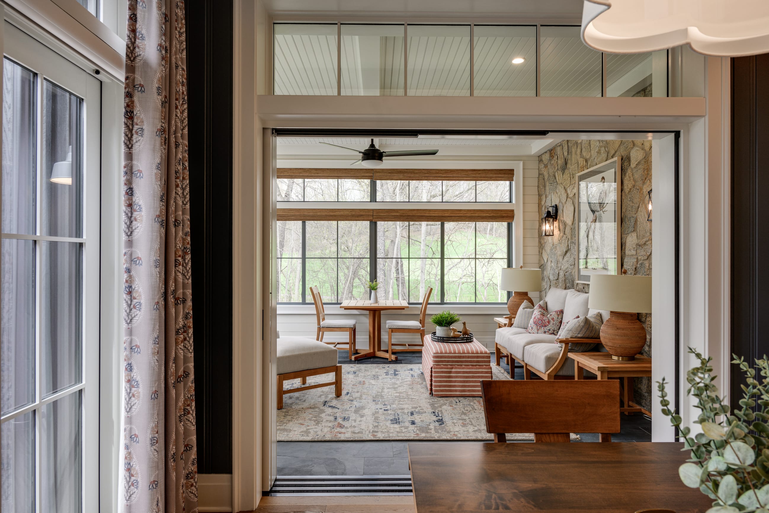 nterior view looking into a bright sunroom or enclosed porch with large windows overlooking a green yard and leafless trees. The space features a small wooden dining table with two chairs, a cushioned bench-style sofa against a stone accent wall, a patterned ottoman, and a light-colored area rug. Warm wood furniture, table lamps, and a ceiling fan add to the cozy atmosphere, while sliding glass doors connect the room to the main interior space.