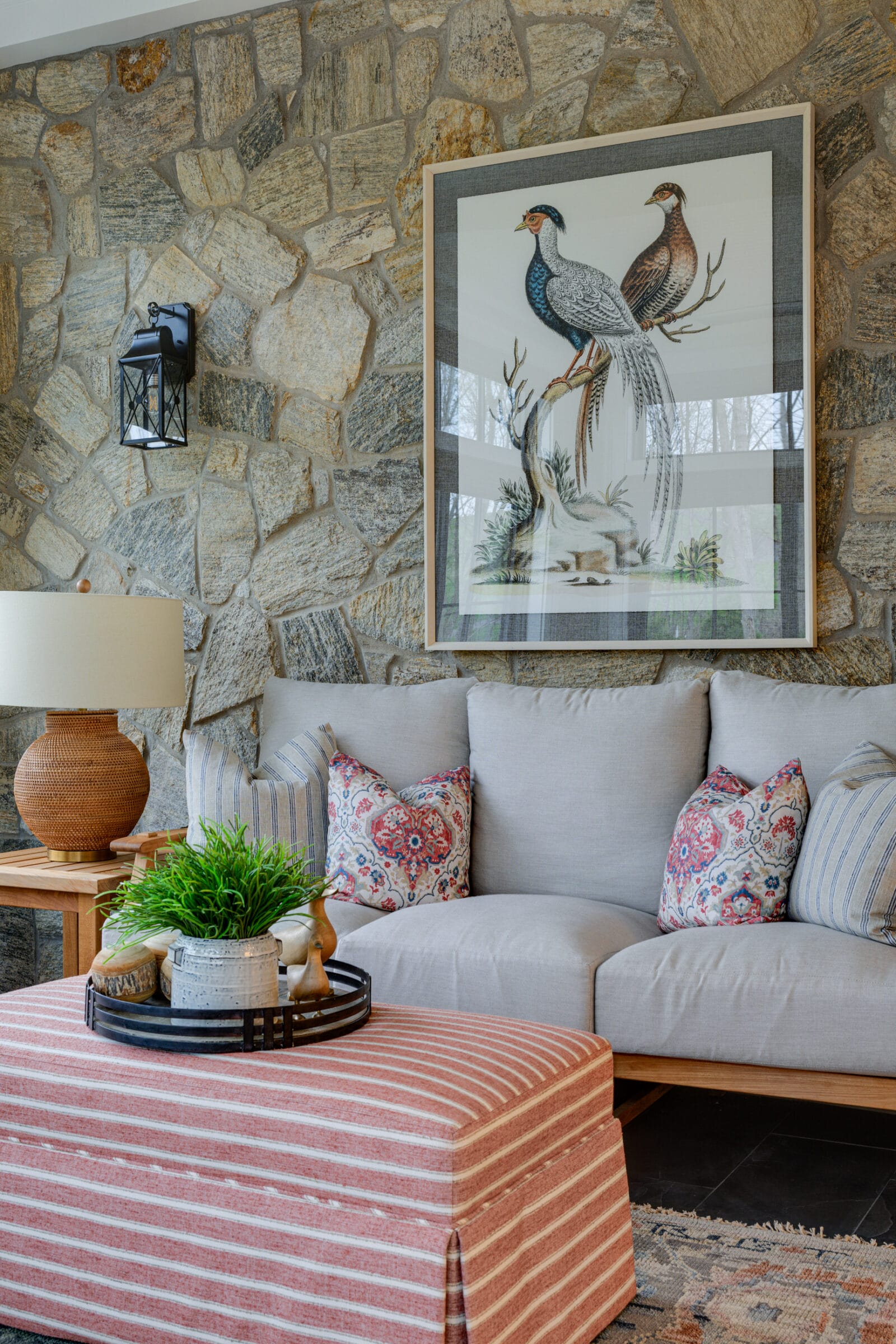 Close-up view of a cozy seating area against a rustic stone wall, featuring a light-colored upholstered sofa with patterned accent pillows. Above the sofa hangs a large framed artwork depicting two birds perched on a branch. A woven table lamp sits on a wooden side table, while a striped upholstered ottoman and a small tray with a plant and decorative accents occupy the foreground. The space combines natural textures and warm tones for a comfortable, inviting interior.