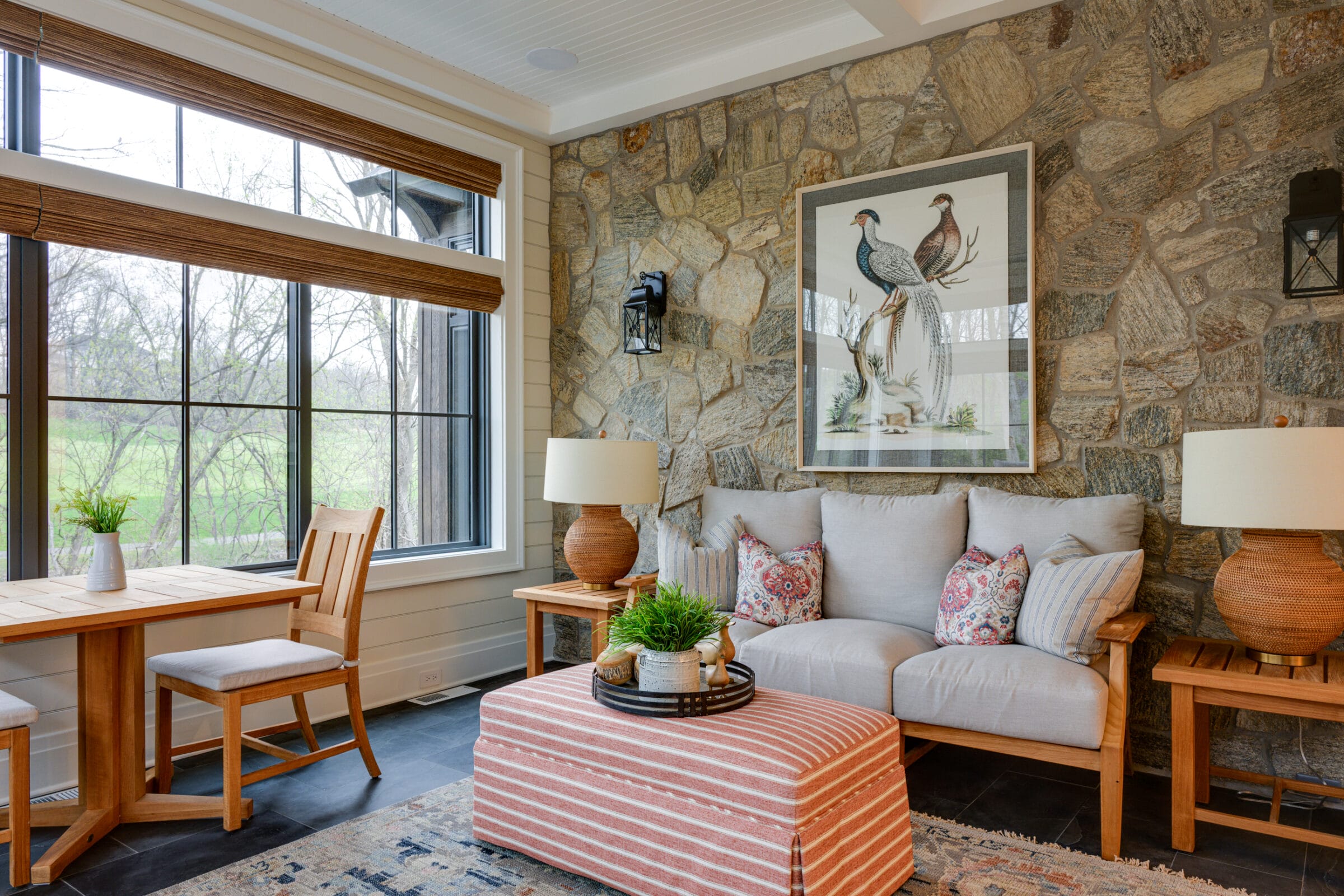 Bright, inviting sunroom with a mix of stone and light-colored walls, featuring a large window with woven wood shades overlooking a green yard and trees. The room includes a wooden dining table with two chairs on the left and a cozy seating area on the right with a light upholstered sofa, patterned accent pillows, and a striped ottoman topped with a decorative tray and plant. A large framed bird illustration hangs on the stone wall between two wall sconces, while matching table lamps on wooden side tables add warmth to the space.