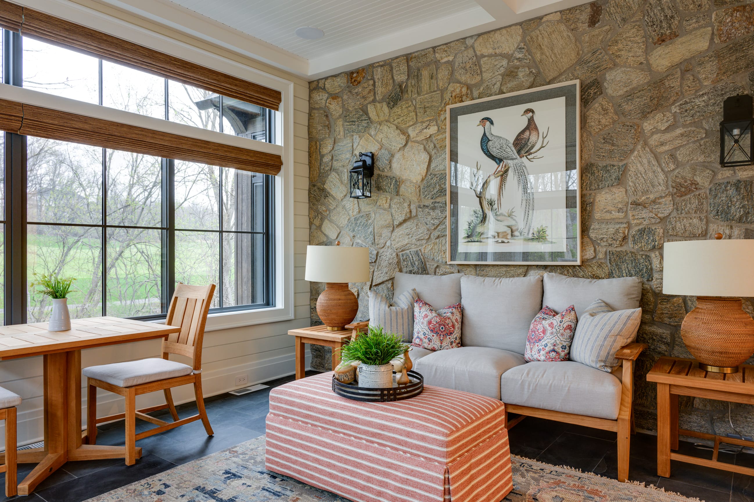 Bright, inviting sunroom with a mix of stone and light-colored walls, featuring a large window with woven wood shades overlooking a green yard and trees. The room includes a wooden dining table with two chairs on the left and a cozy seating area on the right with a light upholstered sofa, patterned accent pillows, and a striped ottoman topped with a decorative tray and plant. A large framed bird illustration hangs on the stone wall between two wall sconces, while matching table lamps on wooden side tables add warmth to the space.