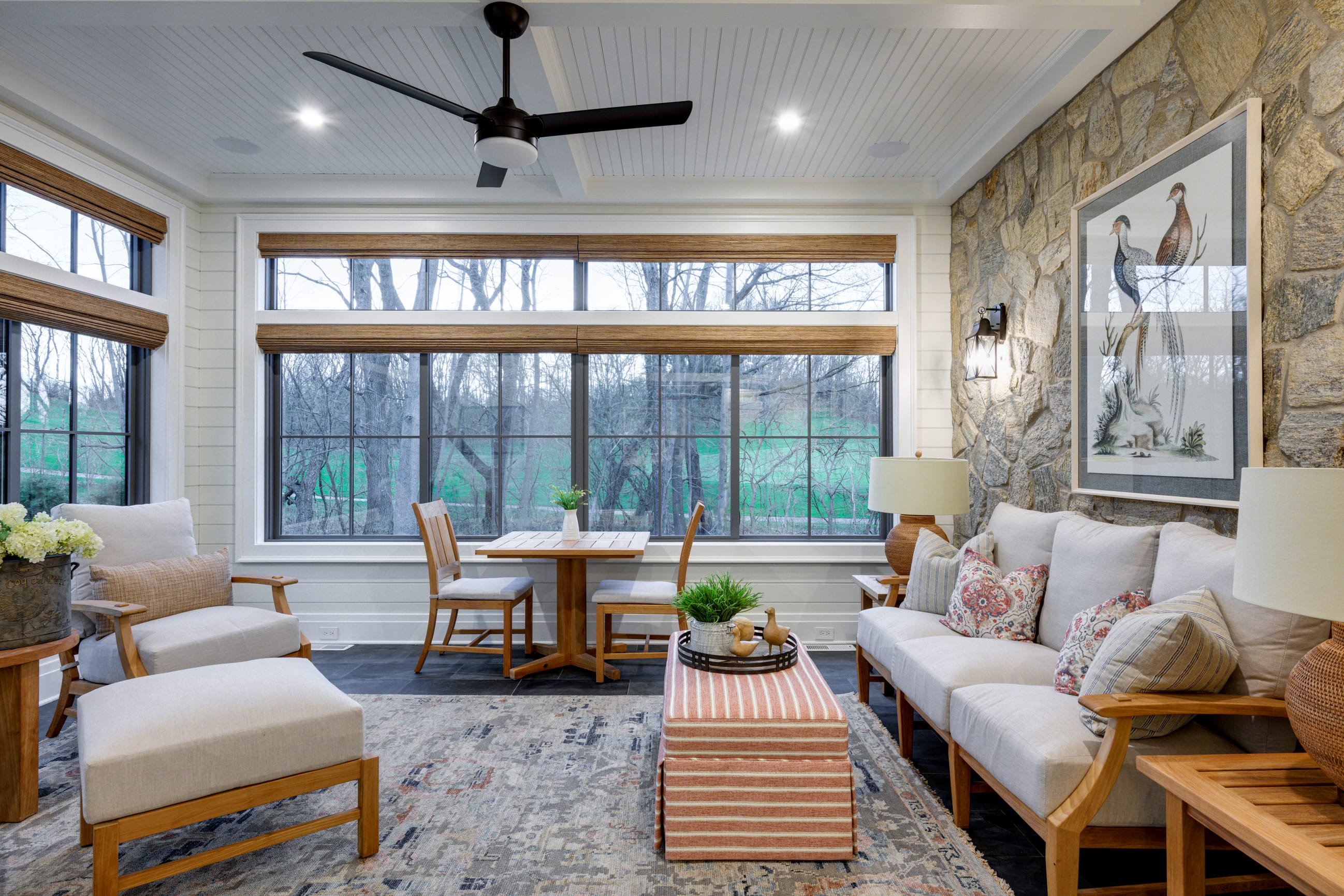 Bright sunroom featuring a stone wall, cozy seating, a wooden table, and large windows overlooking a green landscape.