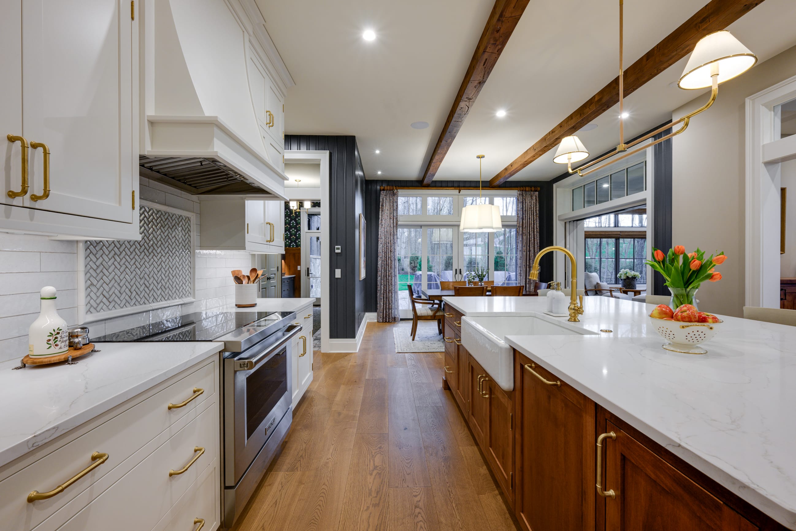 Modern kitchen interior featuring a long central island with a white countertop, a farmhouse-style sink, and gold-toned fixtures. White cabinetry with brass handles lines the left side alongside a stainless steel range and a patterned tile backsplash, while warm wood cabinetry appears on the island. Exposed wooden ceiling beams and recessed lighting add warmth, and pendant lights hang above the island. In the background, a dining area with a wooden table and large windows overlooks a green backyard, connecting the kitchen to an adjacent seating space.