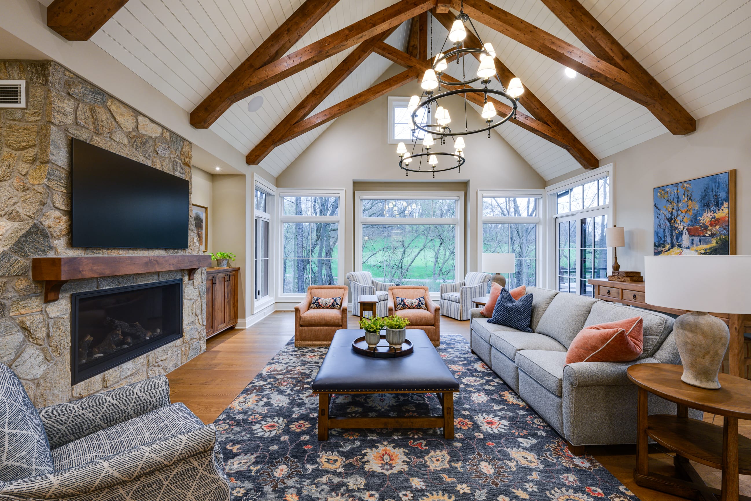 Spacious living room with vaulted ceilings and exposed wooden beams, featuring a large stone fireplace with a mounted flat-screen TV. The room includes a gray upholstered sofa, patterned armchairs, and a leather-topped coffee table arranged on a decorative area rug. Two chandeliers hang from the ceiling, and large windows and glass doors along the back wall provide views of a green yard with trees. Wooden furniture, table lamps, and framed artwork complete the warm, modern rustic design.