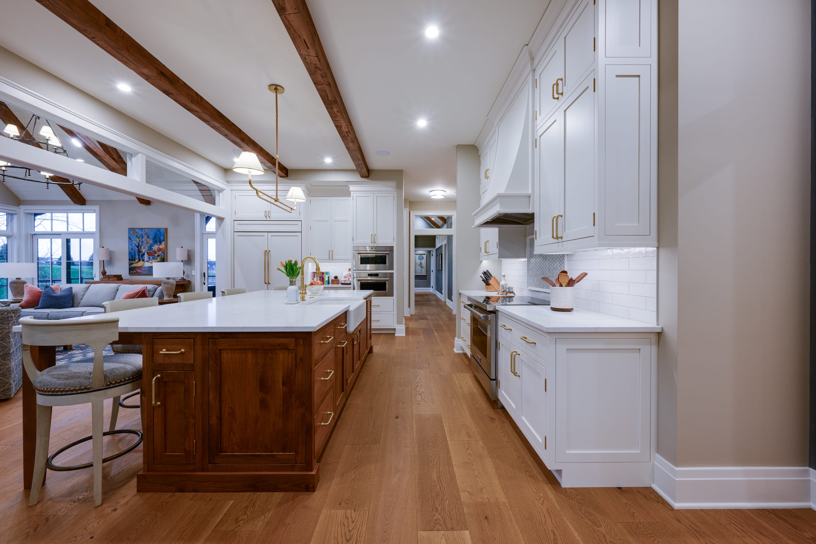 Modern kitchen with white cabinets, wood accents, marble countertops, stainless steel appliances, and exposed ceiling beams. Open layout leads to living area with art on the wall.