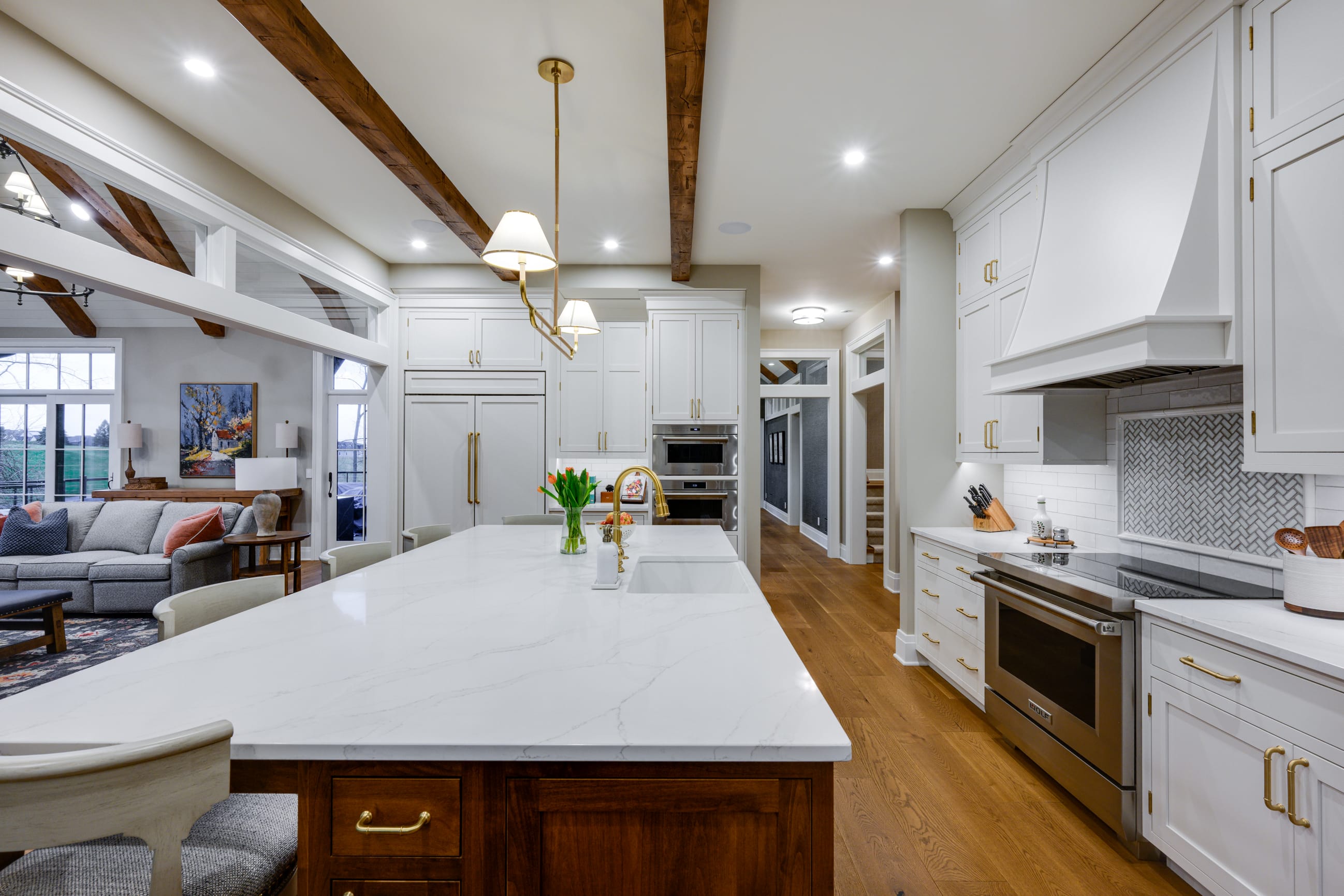 Modern kitchen featuring a large marble island, white cabinetry, wooden beams, and a cozy living area in the background.