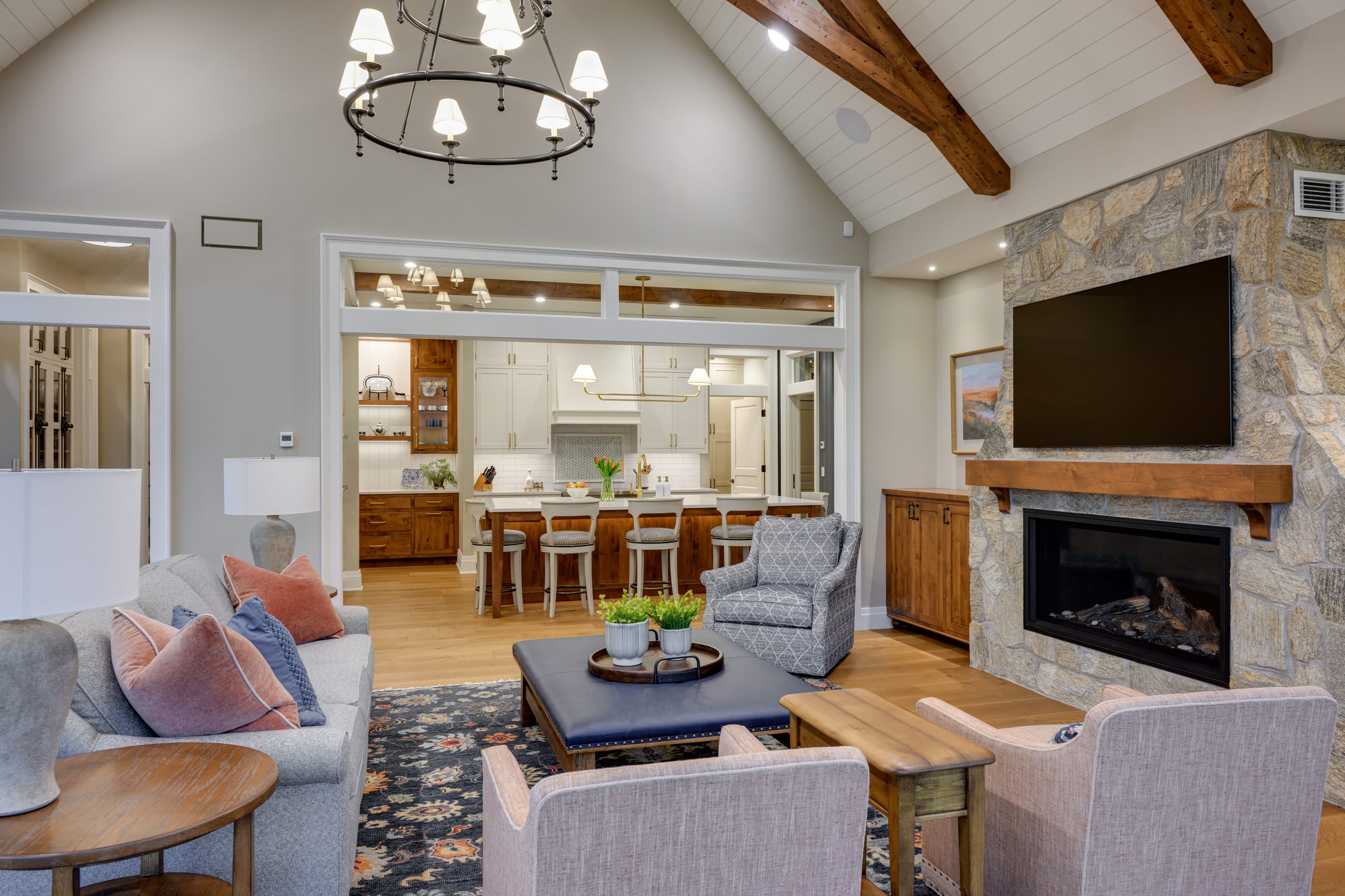 Cozy living room featuring a stone fireplace, modern chandelier, and open kitchen with wooden accents and stylish furnishings.